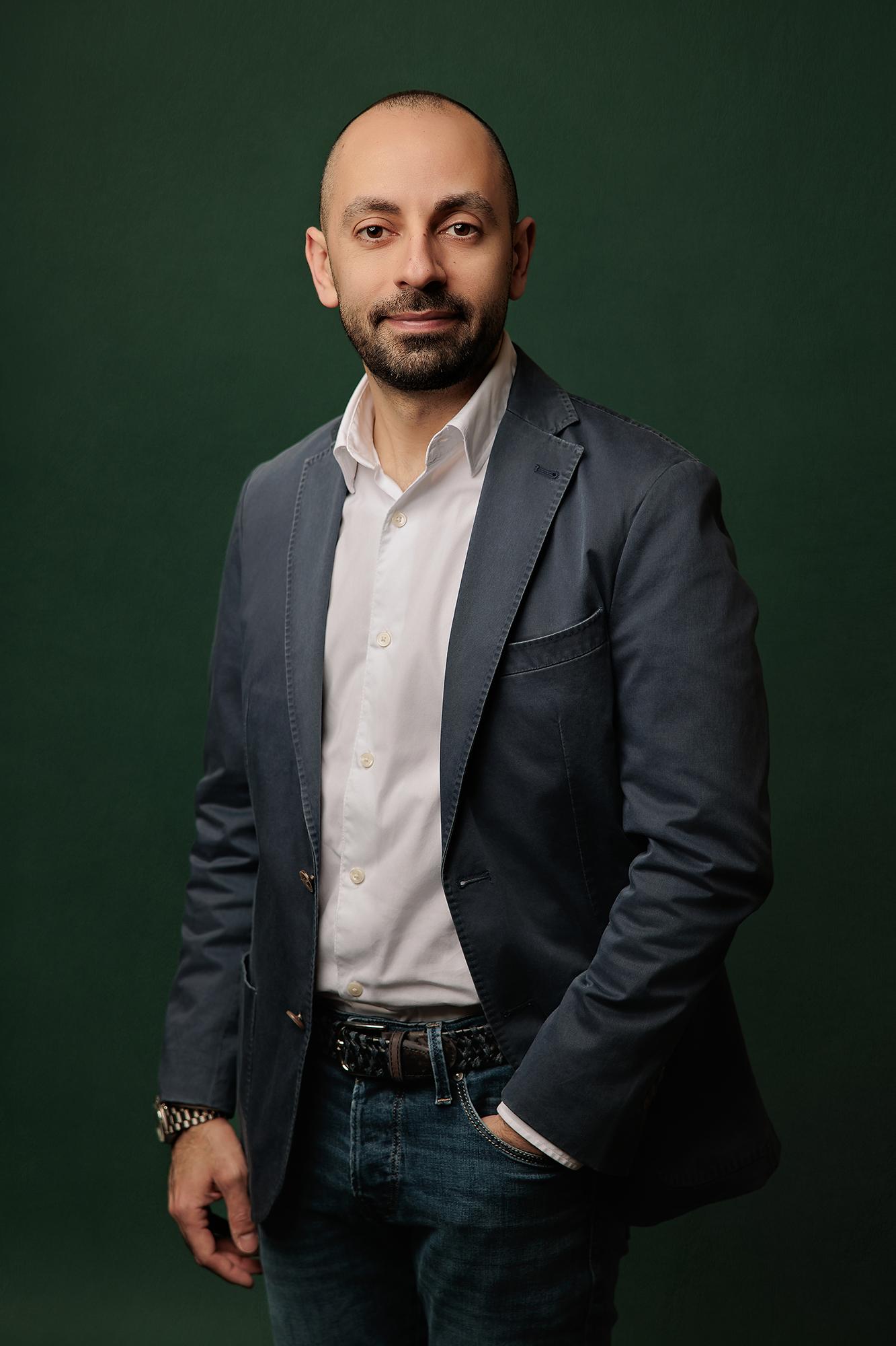A smiling man in a dark blazer and jeans poses for his professional headshot, expertly captured by a Calgary headshot photographer on an emerald green backdrop.