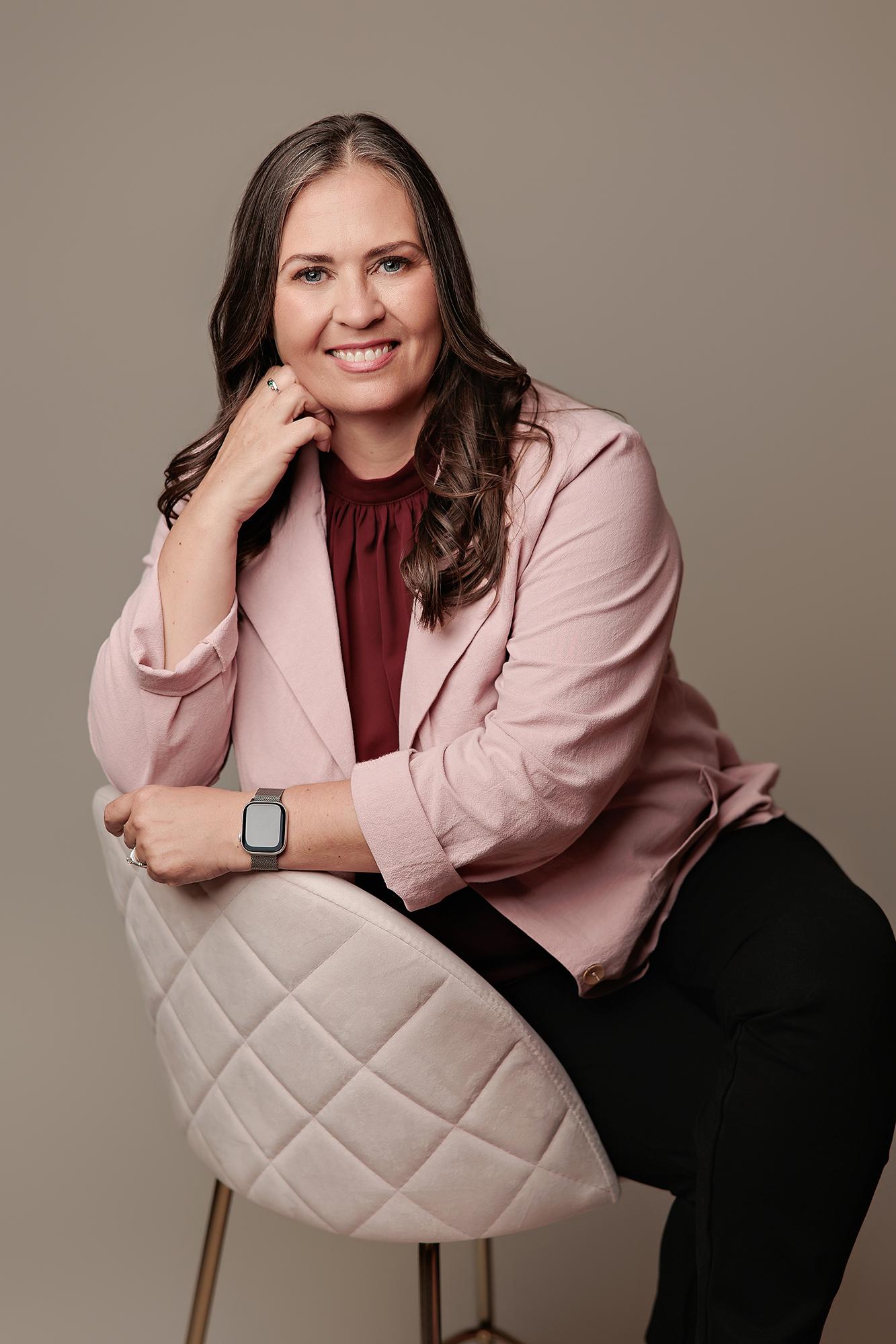 A smiling woman in a pink blazer sits on a chair in a Calgary photography studio, showcasing a professional headshot with soft, flattering lighting.