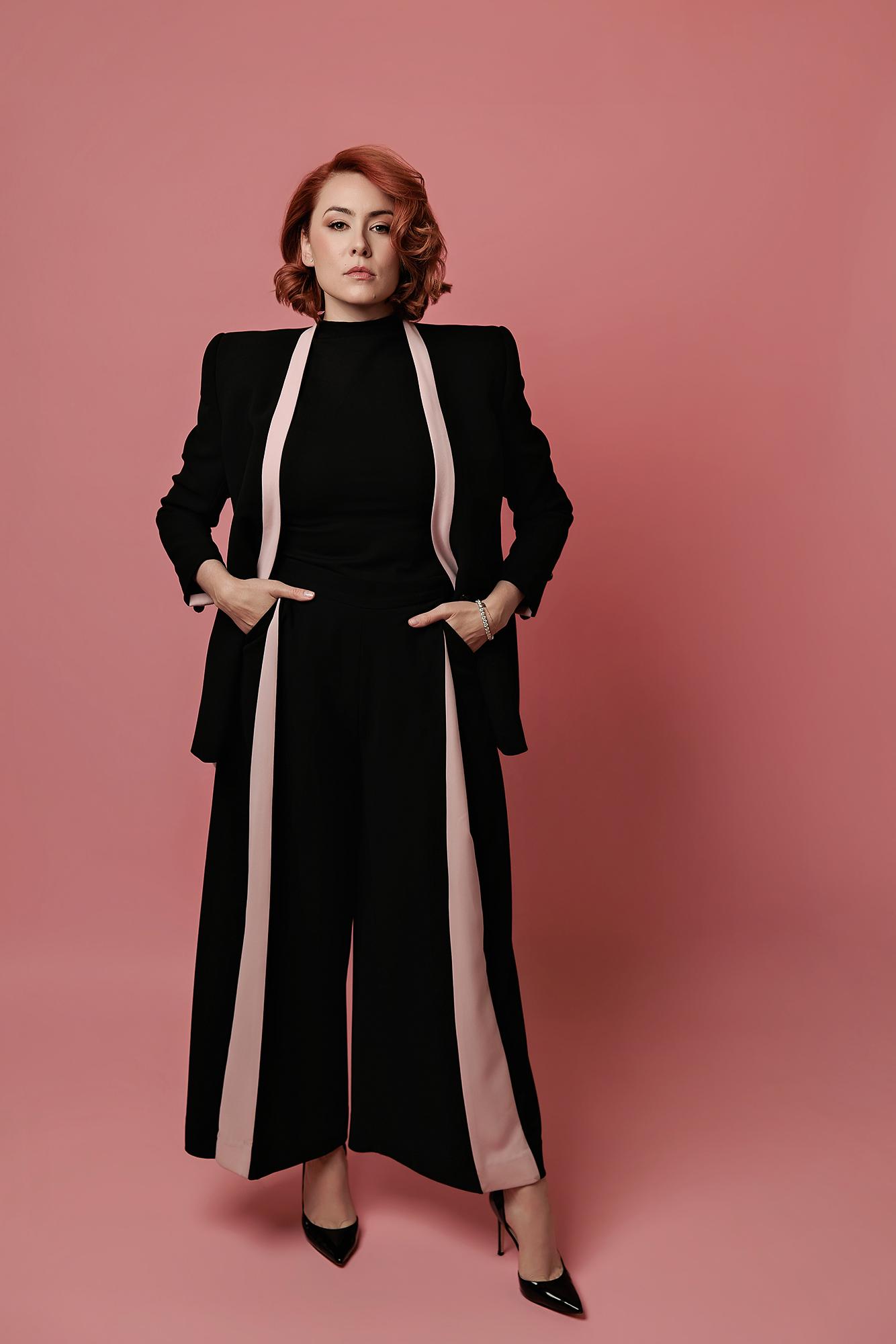 A woman with ginger hair strikes a confident pose in a black suit against a pink backdrop, showcasing professional branding by a Calgary photographer.
