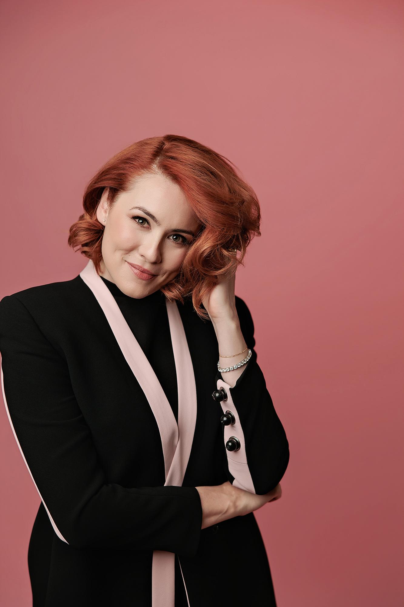A woman with auburn hair poses confidently in a black blazer for a Calgary headshot photography session, set against a soft, professional pink backdrop.