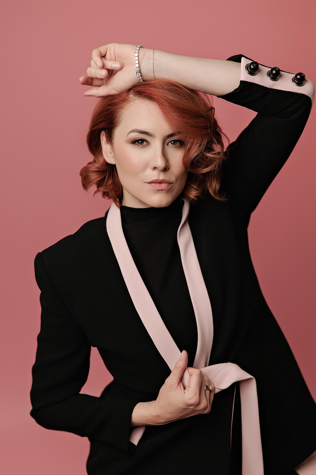 A woman with red hair poses confidently in a black blazer against a pink backdrop, showcasing the bold personal branding style of our Calgary photography studio.