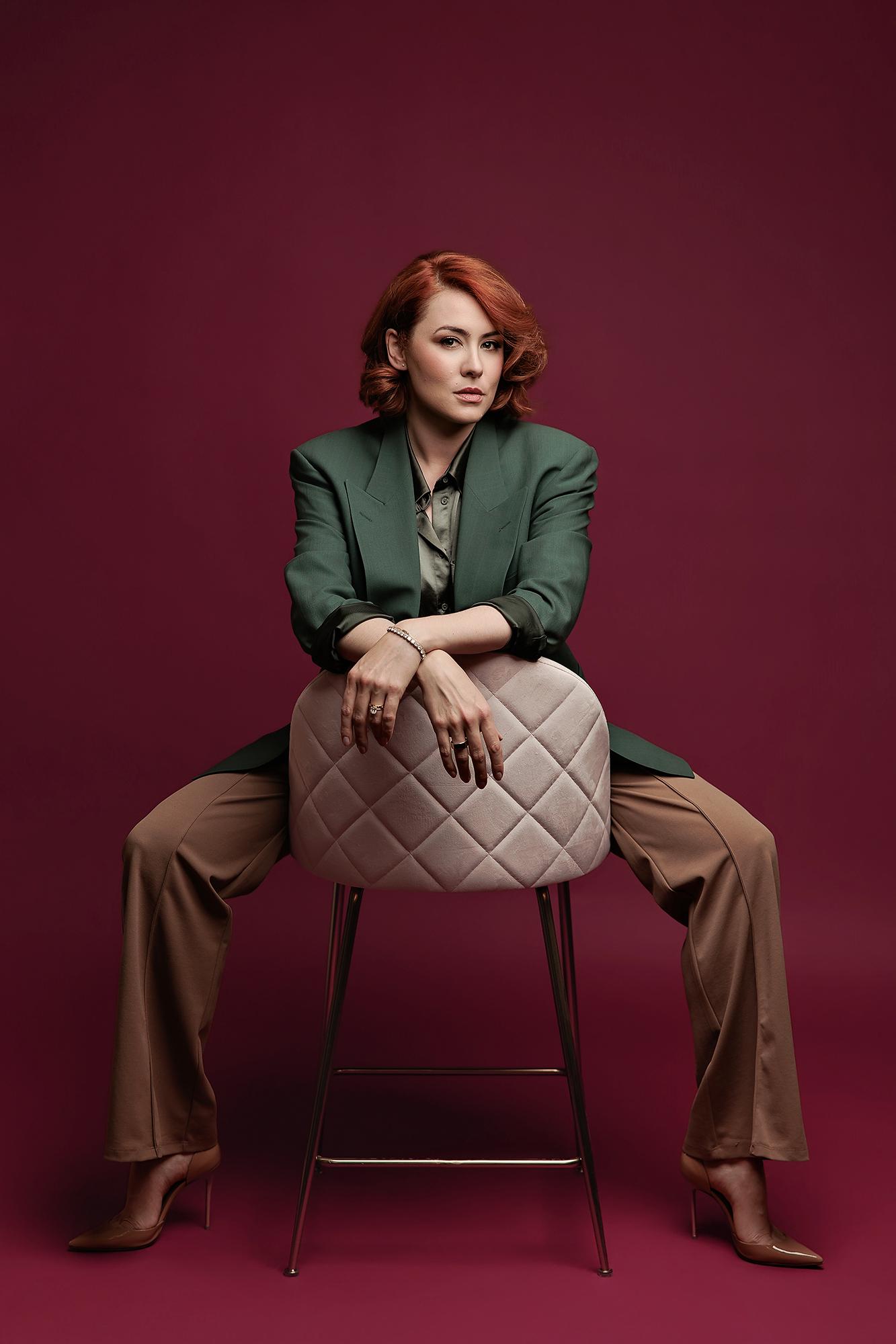 A woman with red hair wears a green blazer and brown trousers while posing on a chair at our Calgary photography studio for a bold personal branding headshot.