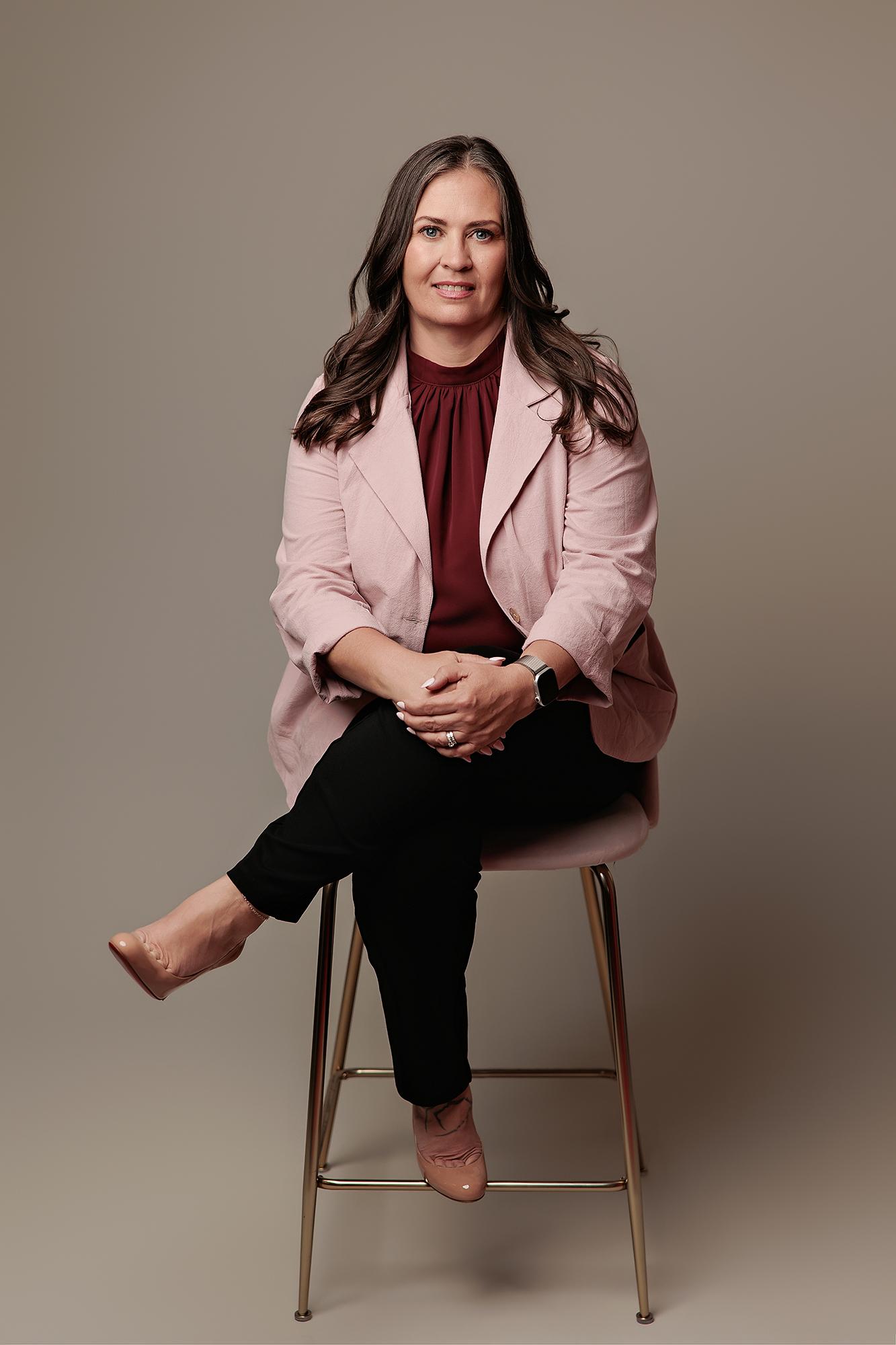 A professional woman in a pink blazer sits on a stool for this Calgary headshot session, showcasing polished studio lighting and a confident pose.