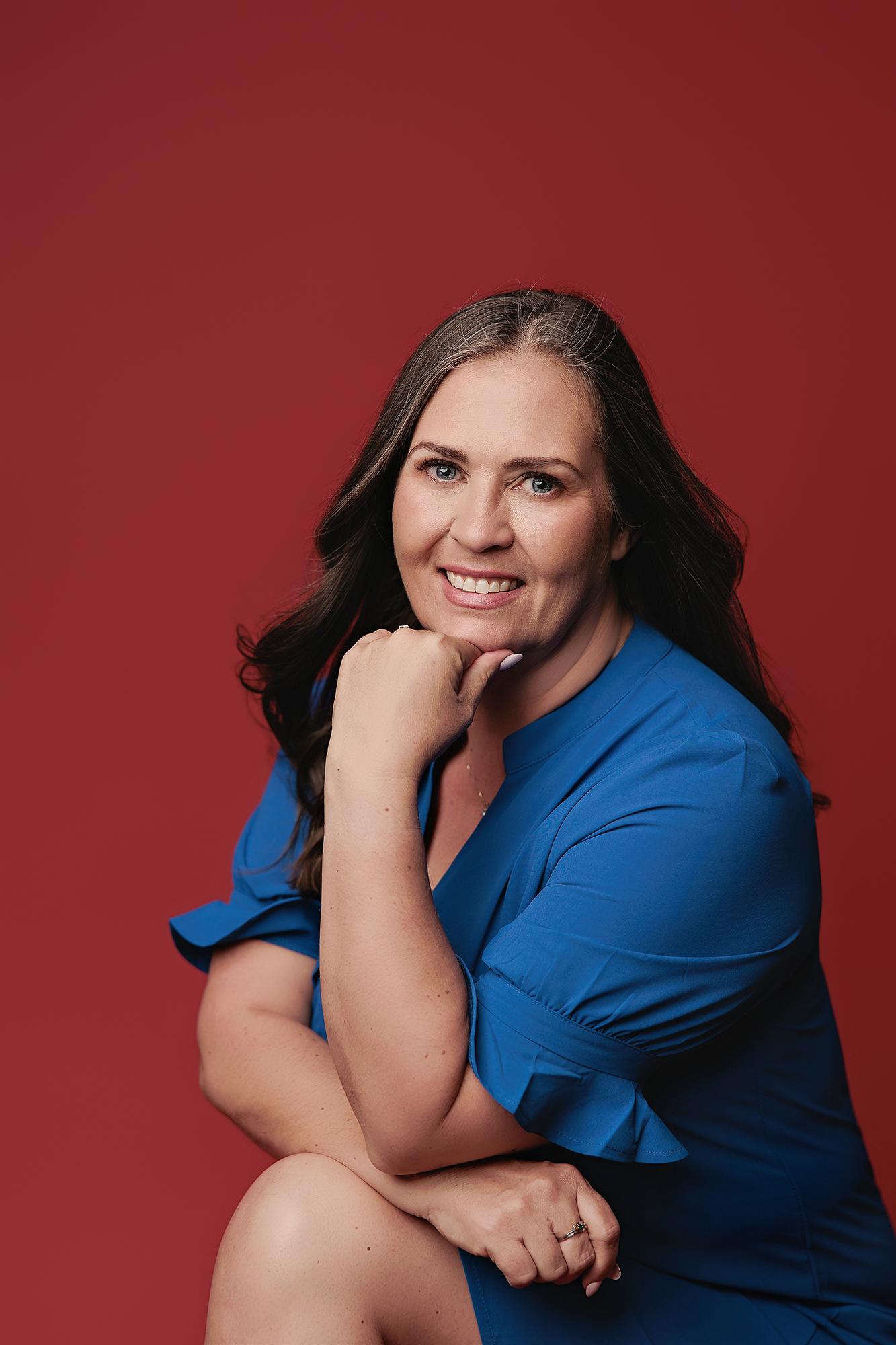 A smiling woman in a vibrant blue dress poses for this Calgary headshot session, showcasing professional lighting against a bold red studio backdrop.