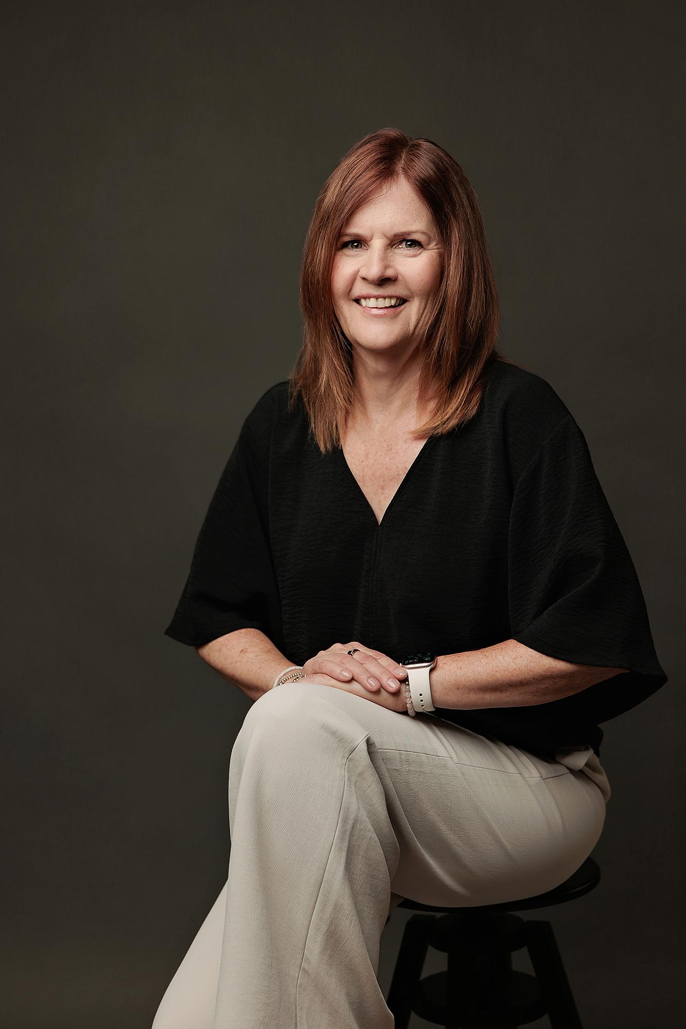 A woman with auburn hair smiles in a professional Calgary headshot studio, wearing a black top and light trousers against a soft, dark backdrop.