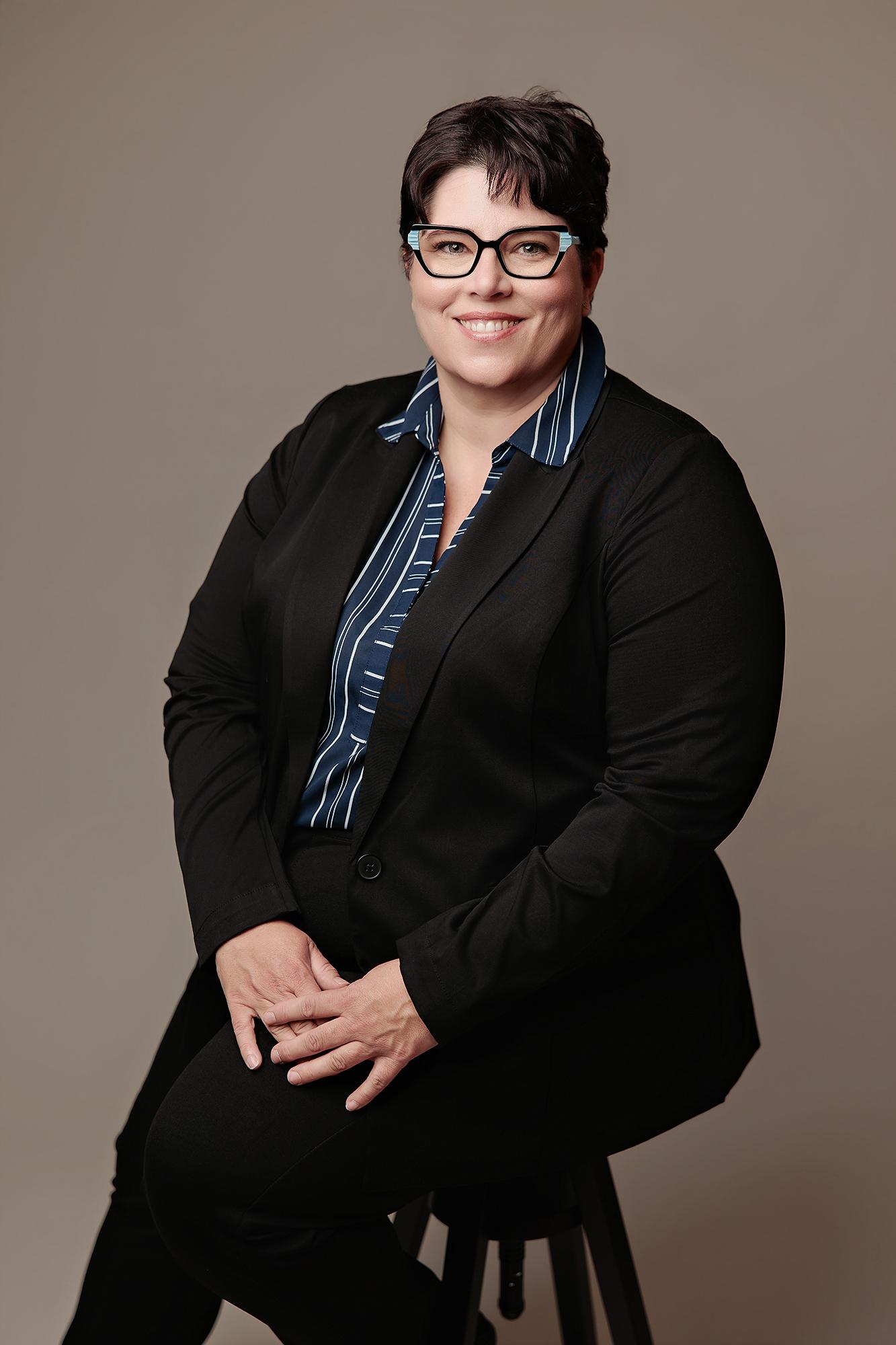 A smiling woman in a black blazer and striped shirt poses for a professional Calgary headshot, captured with soft studio lighting against a neutral backdrop.