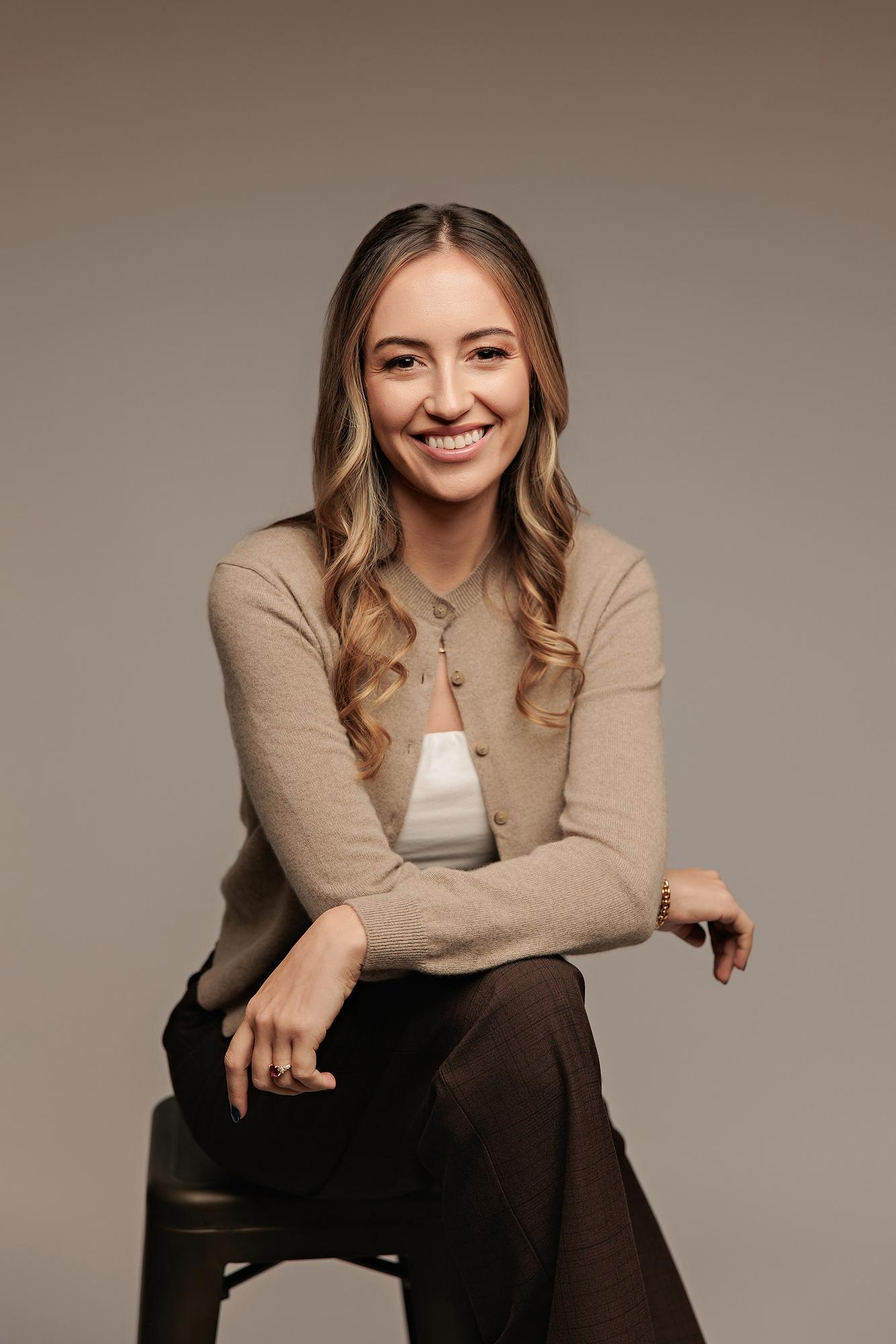 Calgary headshot photographer: Relaxed and approachable young professional woman in a tan cardigan seated on a dark stool against a neutral grey backdrop