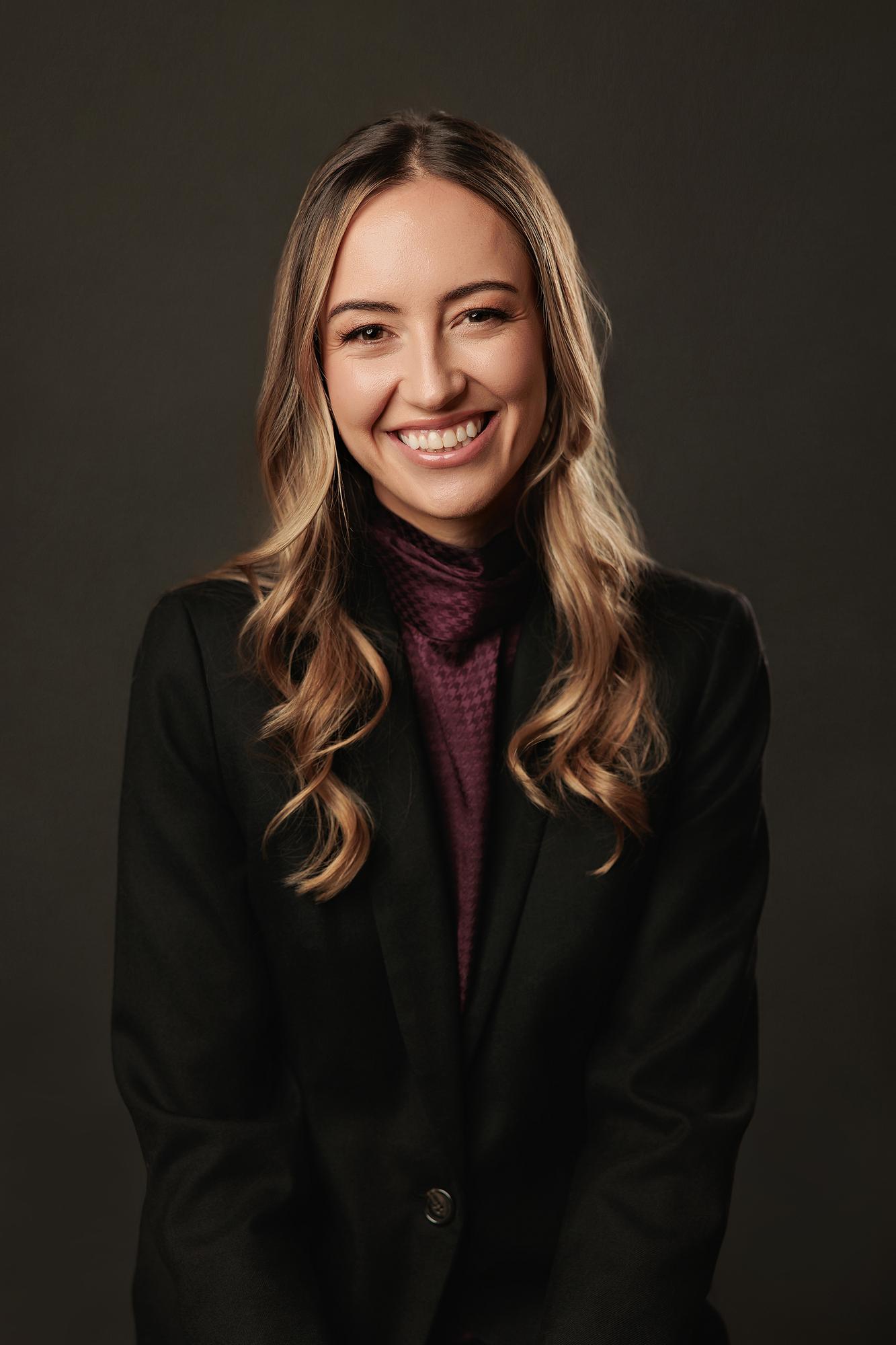 Professional headshot Calgary: Close-up portrait of a smiling young woman in a black blazer and burgundy turtleneck against a dark studio background