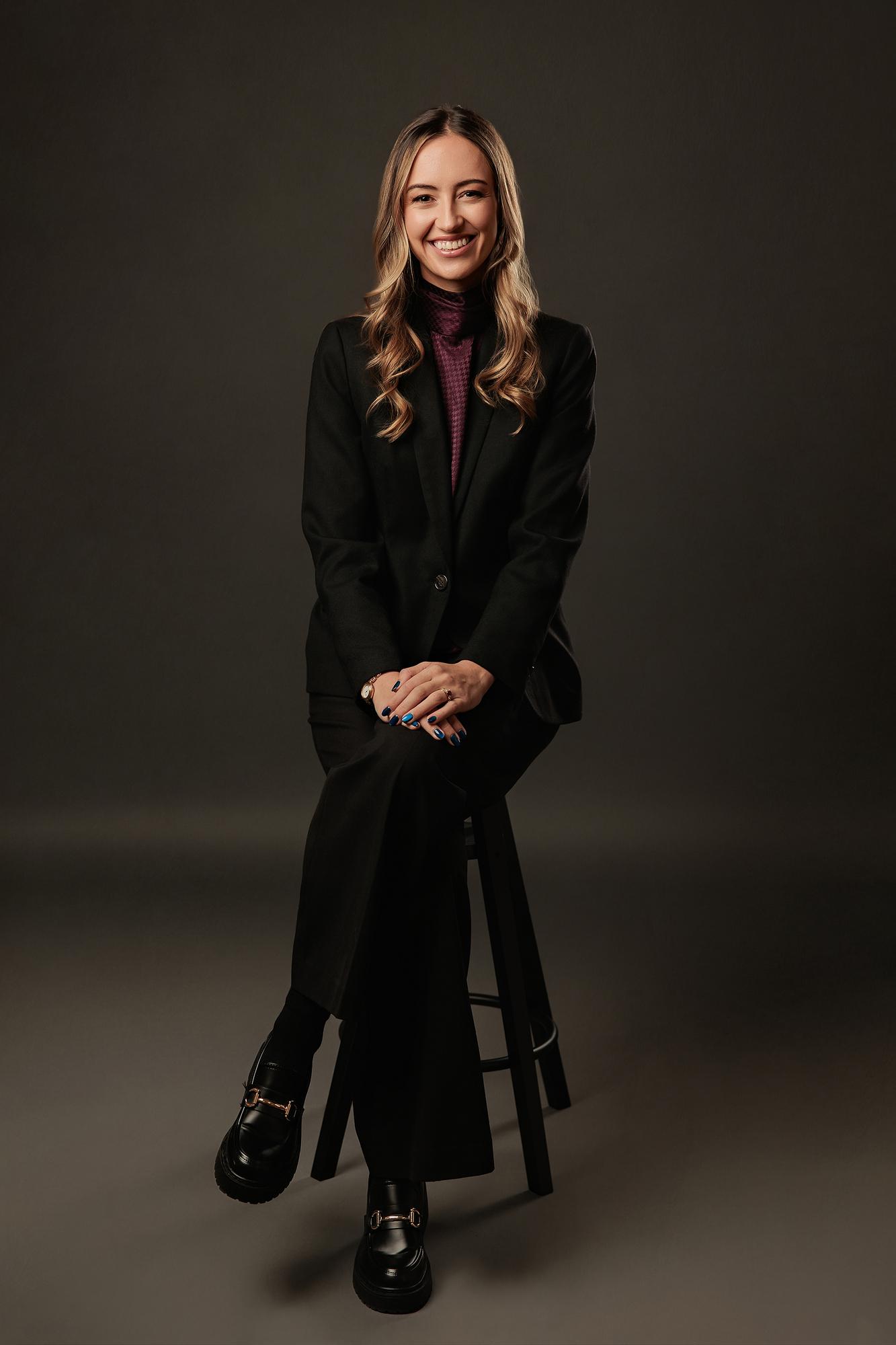 Full-length professional headshot Calgary: Young woman in a black blazer and burgundy turtleneck seated on a dark stool with hands clasped, dark studio background