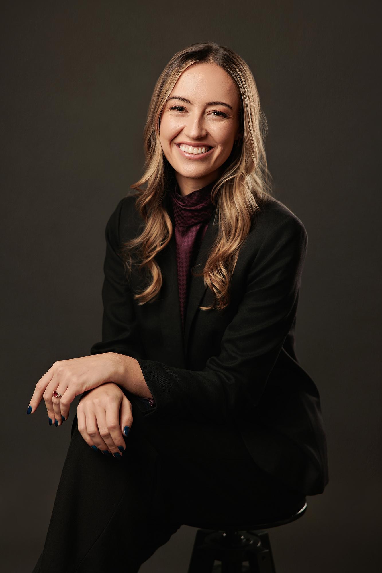 Executive portrait photography Calgary: Smiling young professional woman in a black blazer and burgundy turtleneck leaning forward on a studio stool