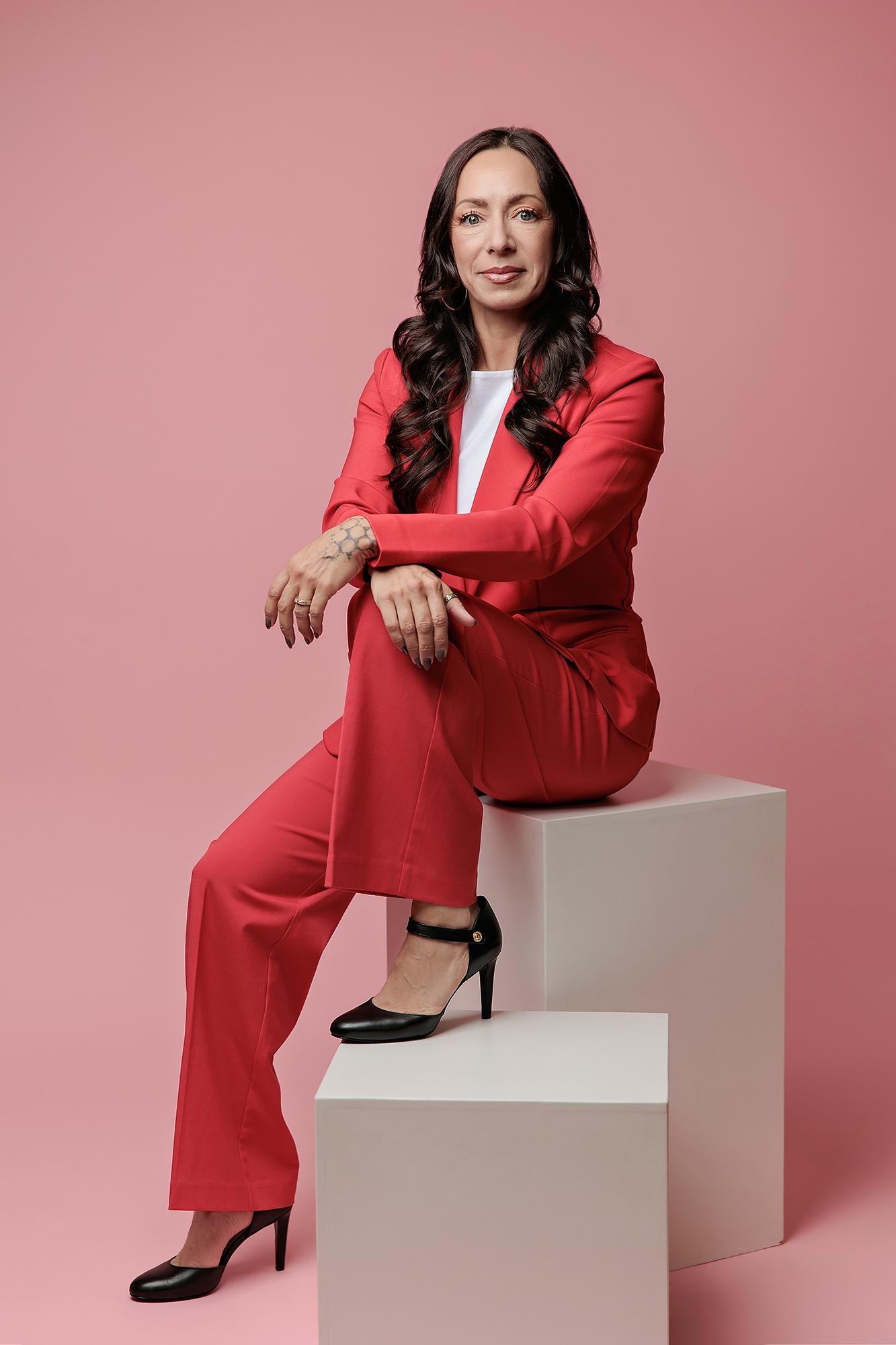 A woman in a vibrant red suit sits confidently on white blocks for a Calgary studio headshot, showcasing professional branding with soft lighting.