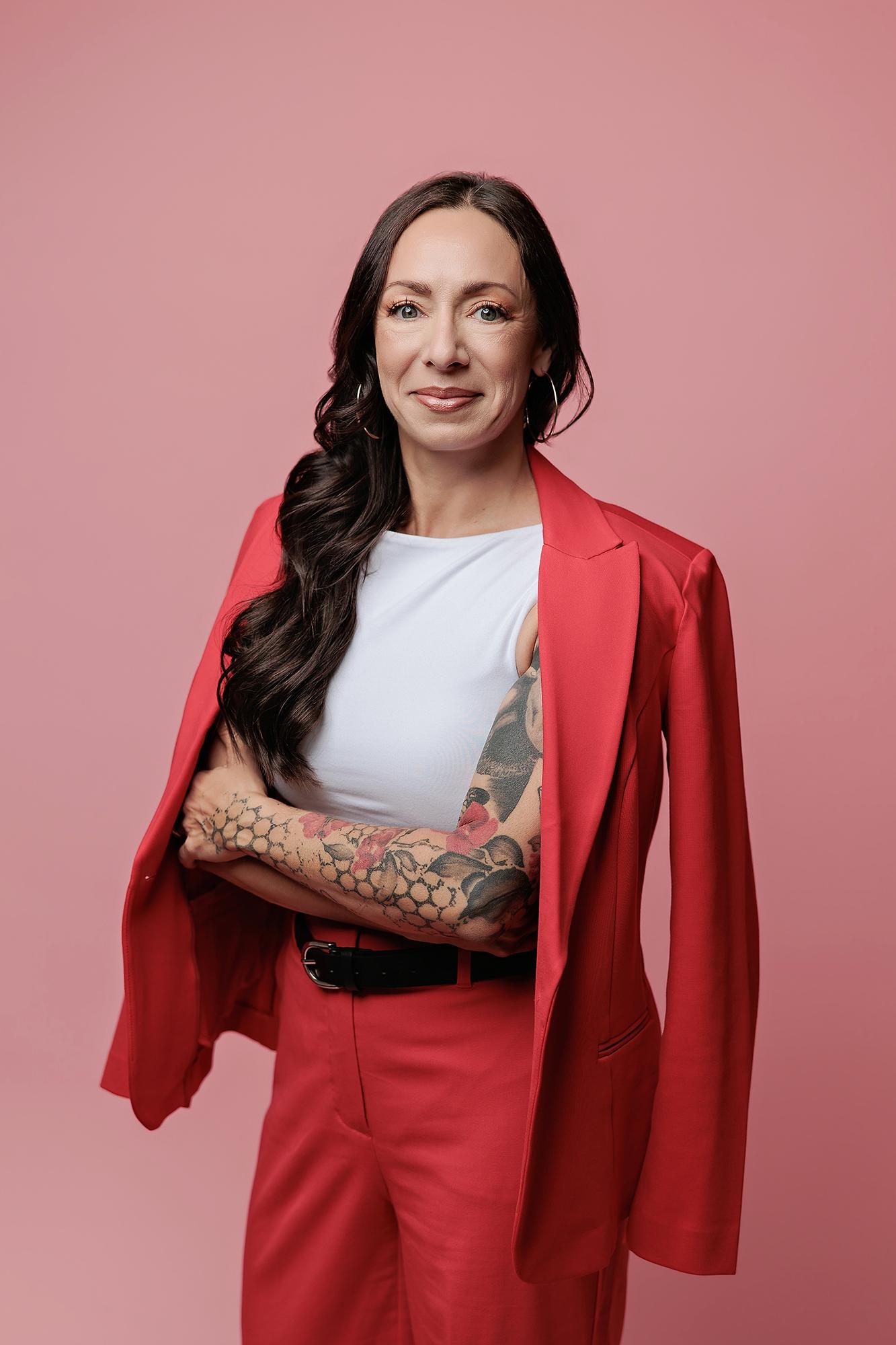 A woman with long wavy hair and arm tattoos wears a red suit in this professional headshot taken at our Calgary photography studio, exuding confidence.