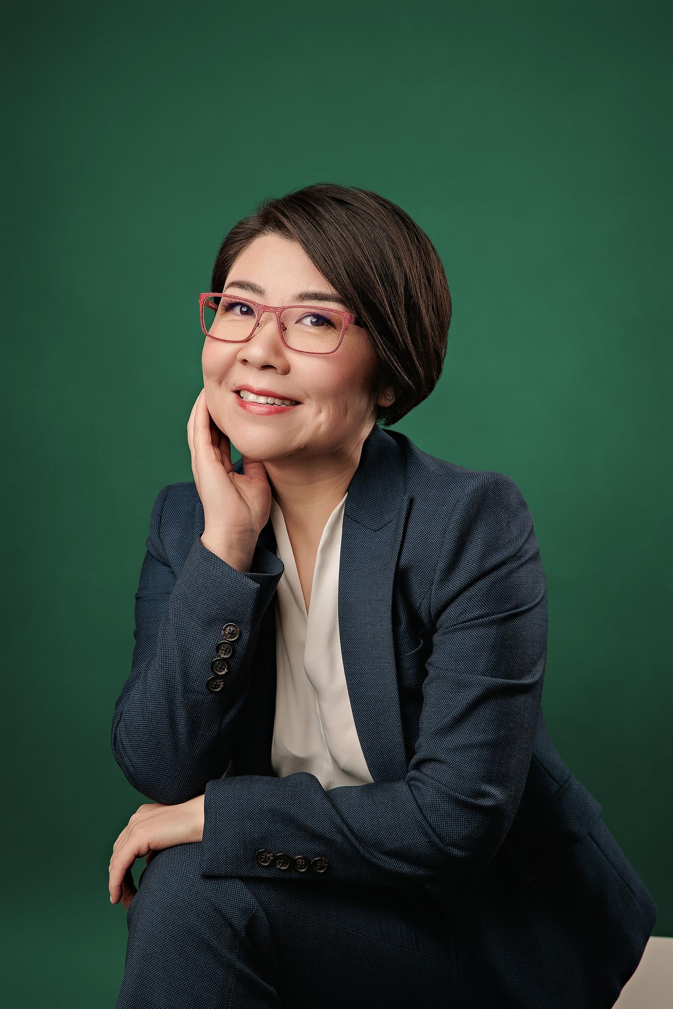 Creative professional headshot Calgary: Woman with red-frame glasses in a navy blazer resting her chin on her hand with a thoughtful expression against a bold green background