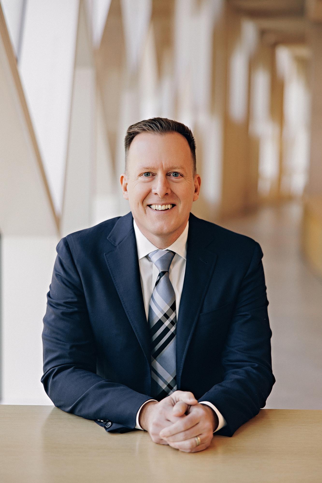 On-location professional headshot Calgary: Smiling man in a navy blazer and plaid tie seated at a table in a bright contemporary office corridor