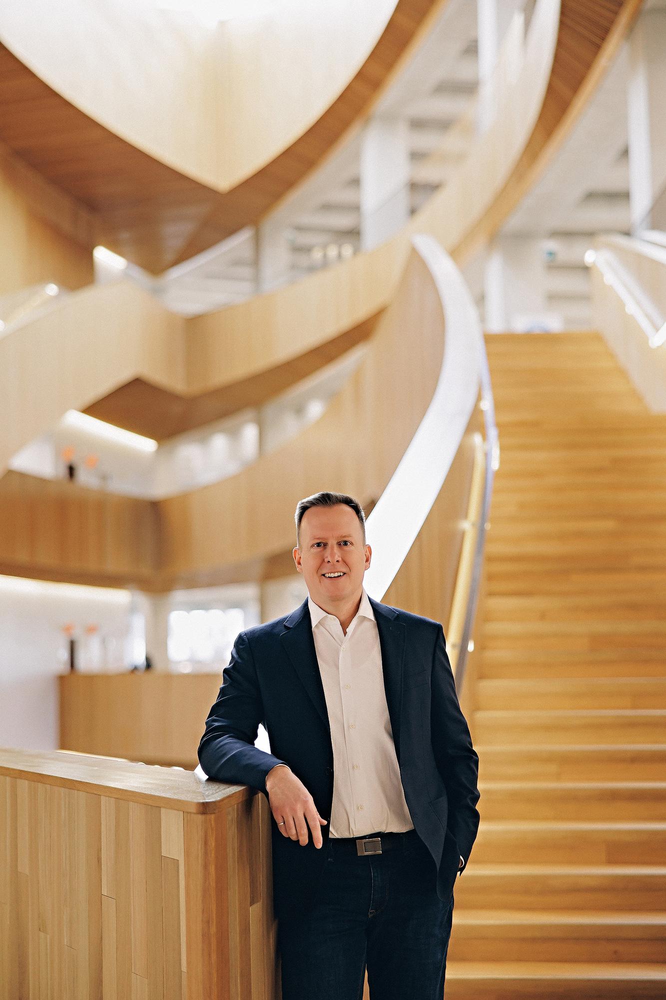 Environmental personal branding headshot Calgary: Professional man in a navy blazer leaning on a wood railing inside a stunning curved architectural staircase atrium