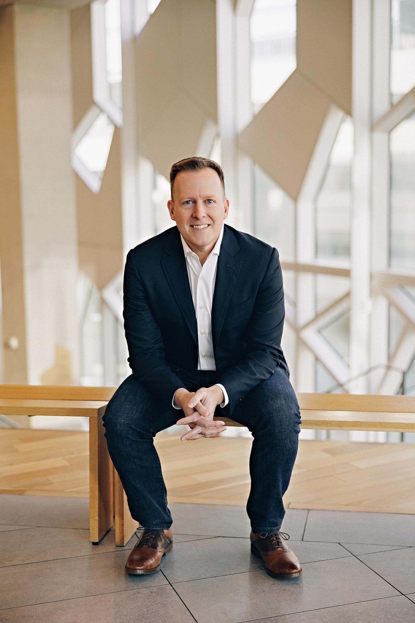 On-location professional headshot Calgary: Smiling man in a navy blazer and white shirt seated on a wood bench in front of a dramatic geometric window wall