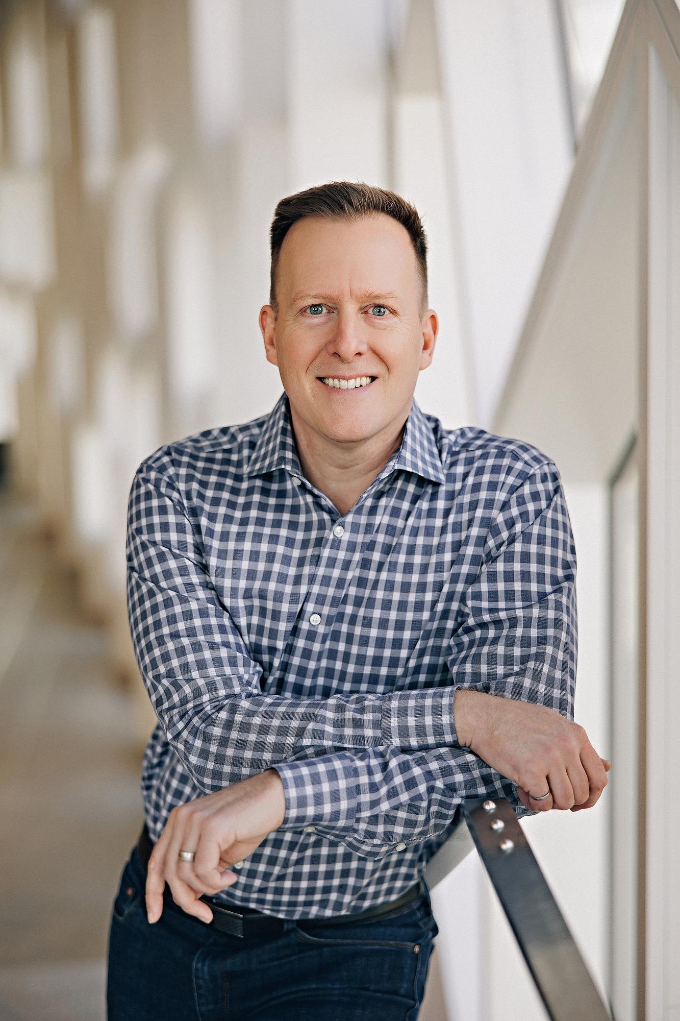 Casual professional headshot Calgary: Approachable man in a blue gingham check shirt leaning on a railing in a bright modern architectural corridor