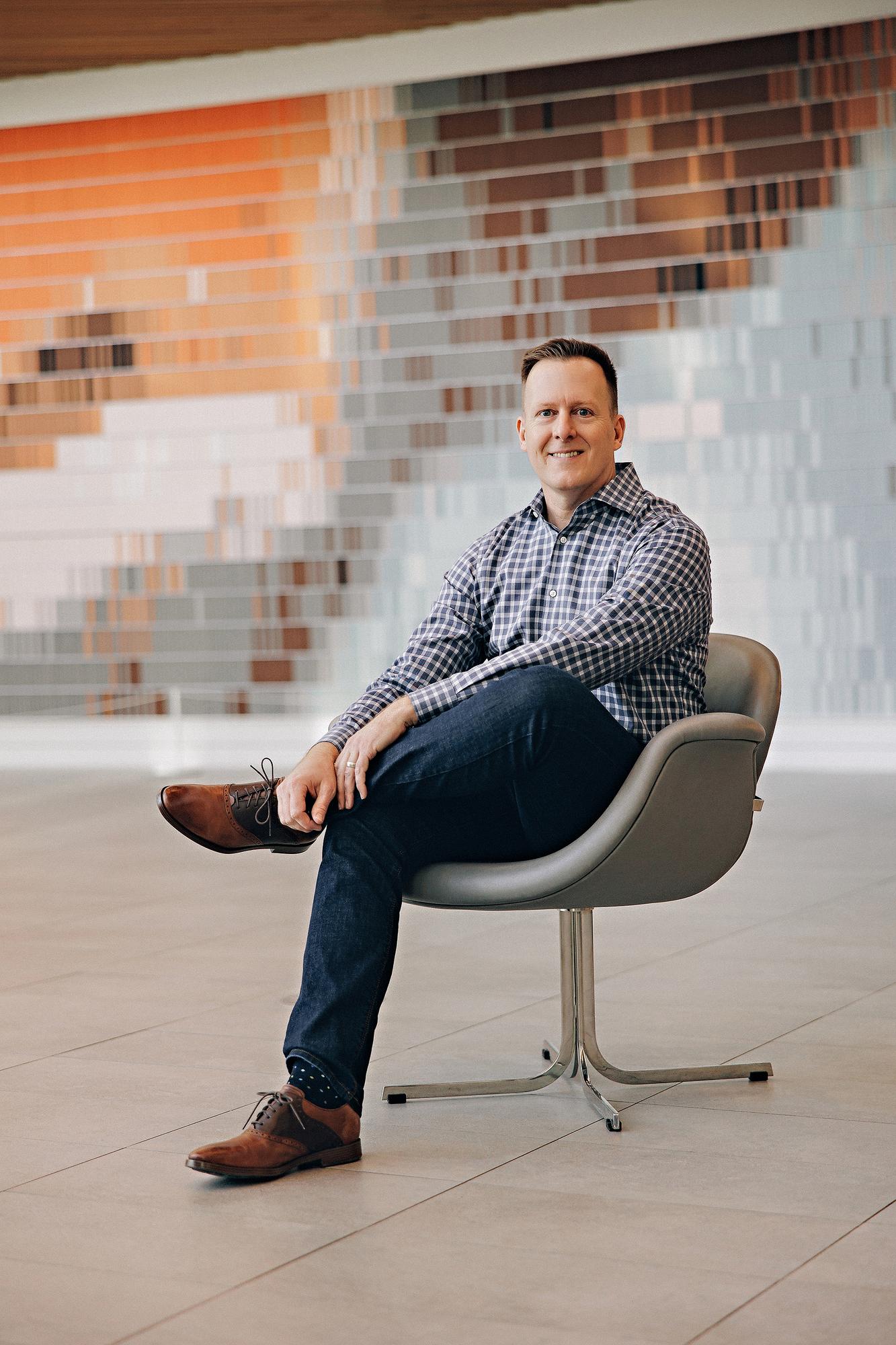 Personal branding photography Calgary: Relaxed professional man in a check shirt seated in a modern lounge chair in front of a colourful mosaic feature wall