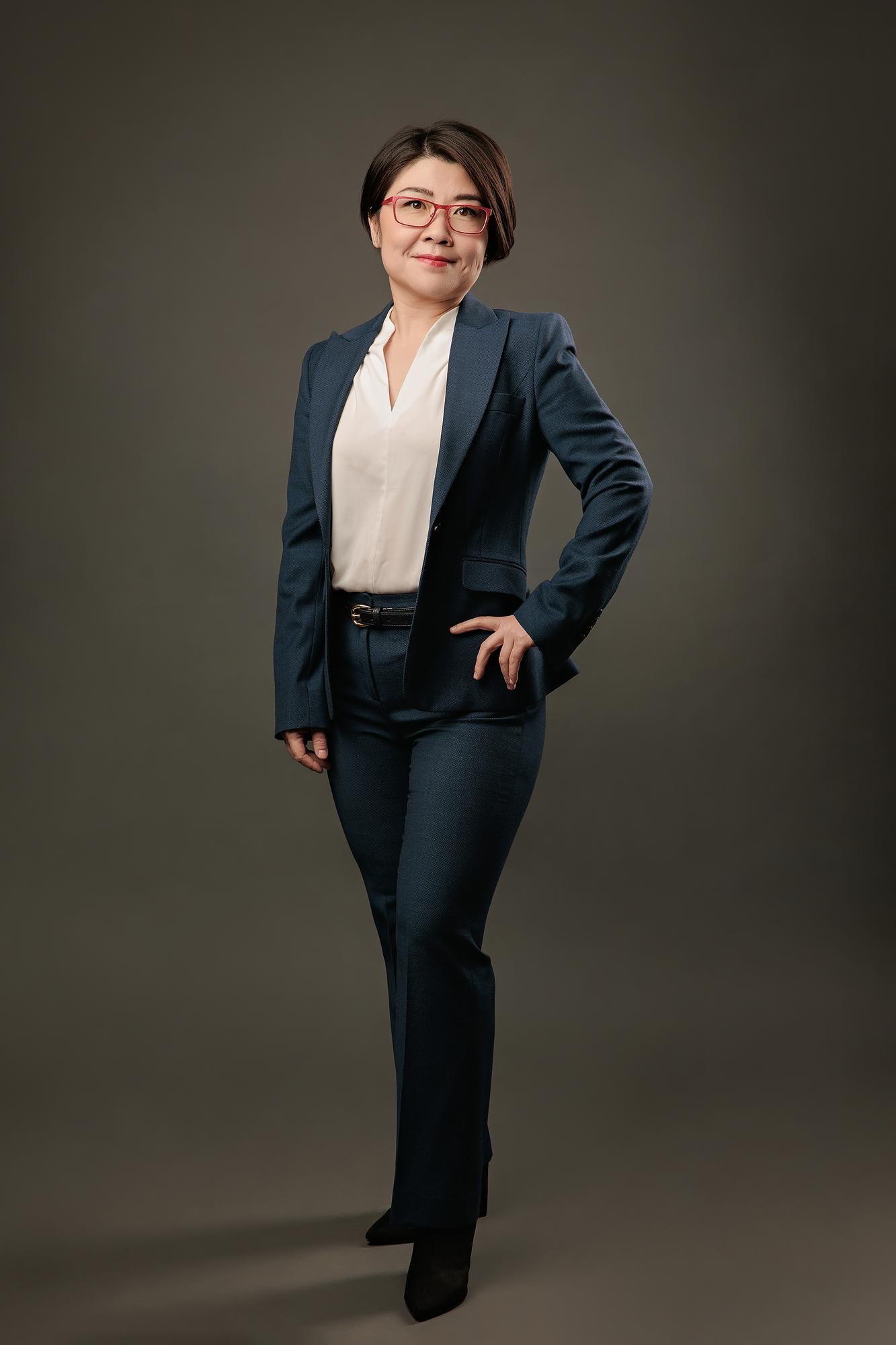 Full-length professional headshot Calgary: Confident woman with red-frame glasses in a teal suit standing with hand on hip against a dark studio background