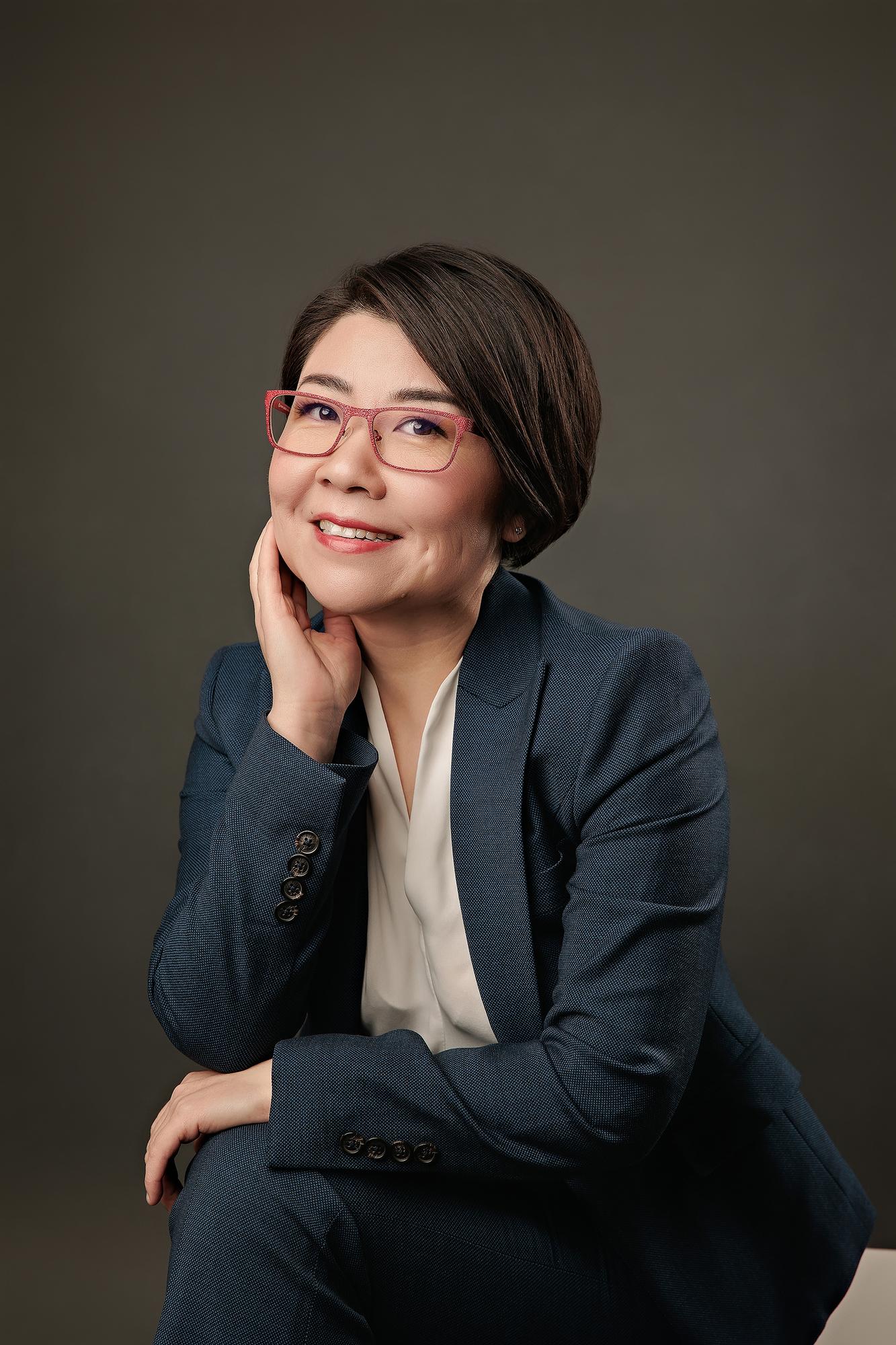 Professional headshot Calgary: Elegant woman with red-frame glasses in a teal blazer and white blouse seated and looking upward with a warm expression, dark background