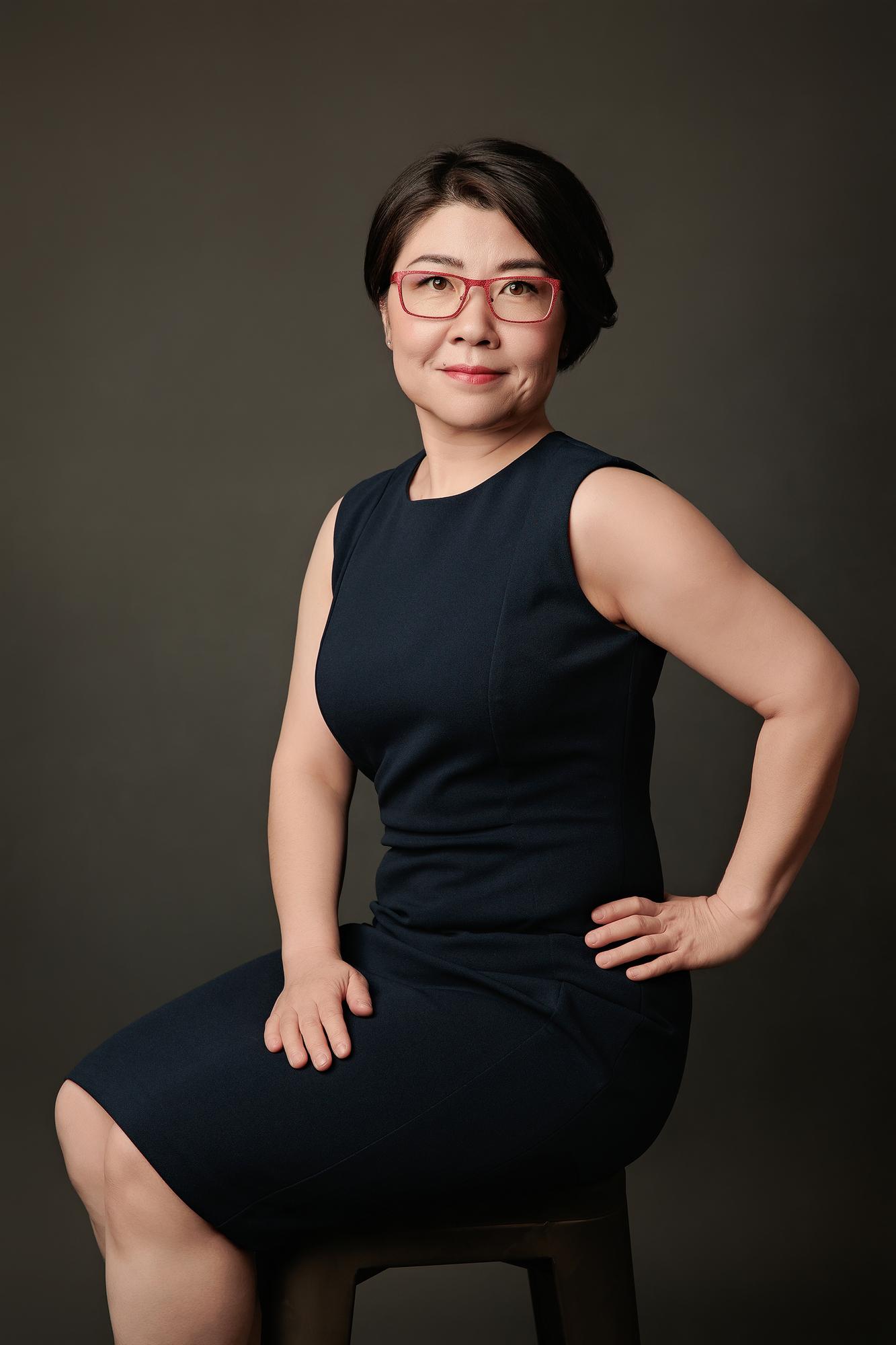Executive portrait photography Calgary: Poised woman with red-frame glasses in a navy sleeveless dress seated on a stool with hand on hip, dark studio background