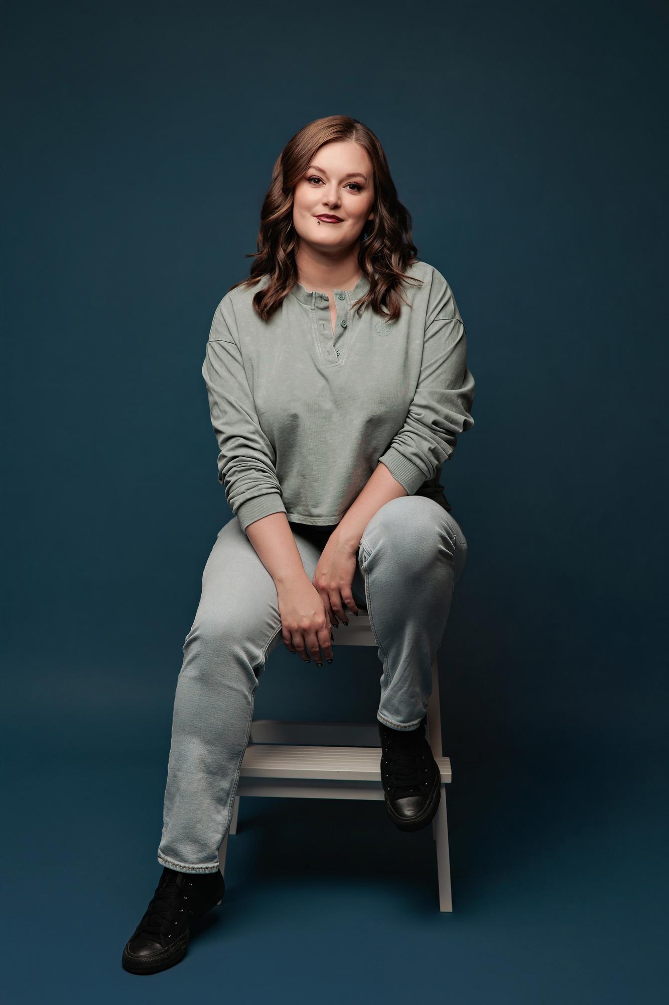 Creative personal branding photography Calgary: Young woman in a sage green Henley and light jeans seated on a white cube with a relaxed expression, teal background