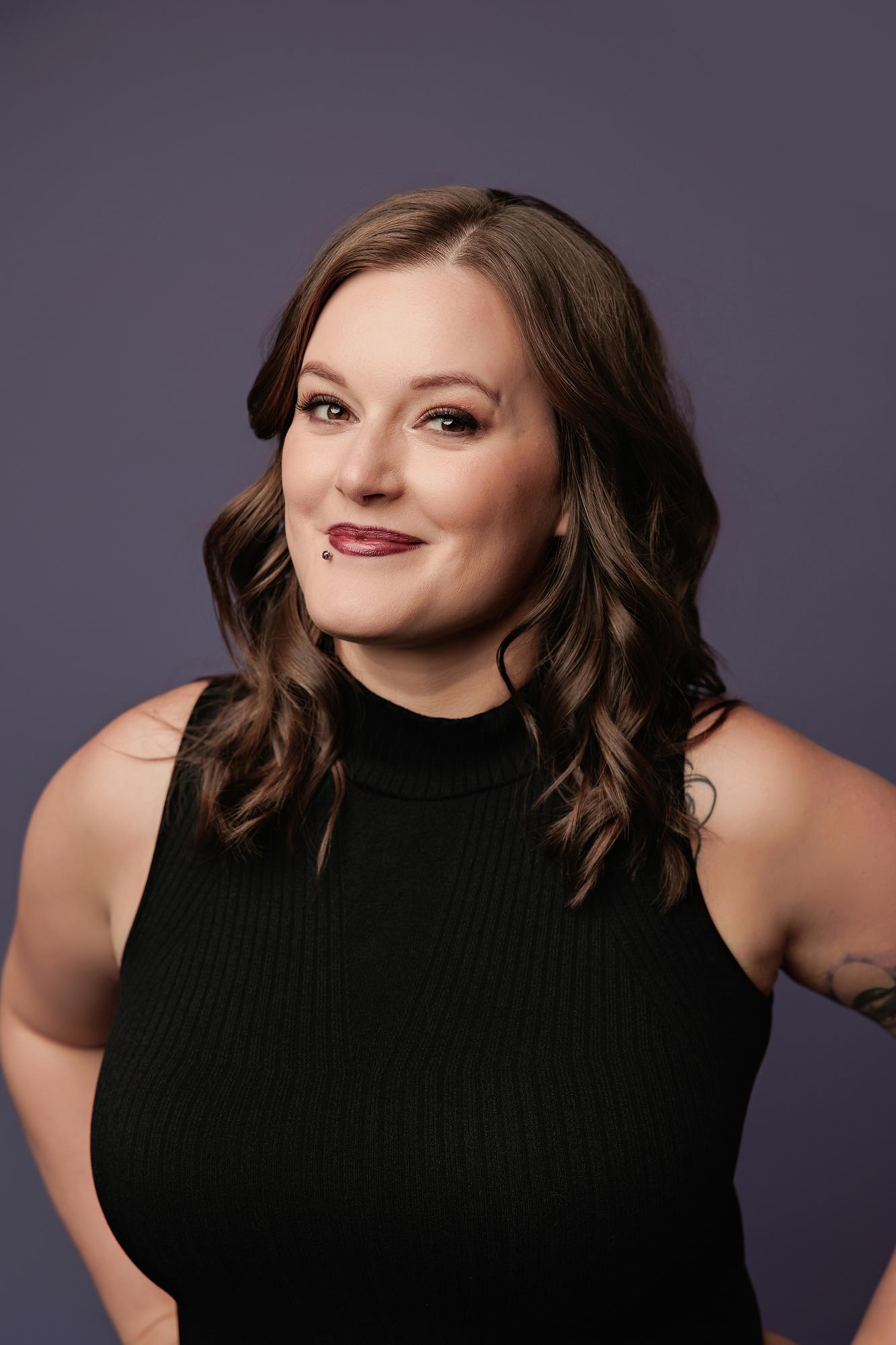 Creative personal branding headshot Calgary: Close-up of a young woman in a black mock-neck top and checker pants with hands on hips and a smouldering expression, purple background