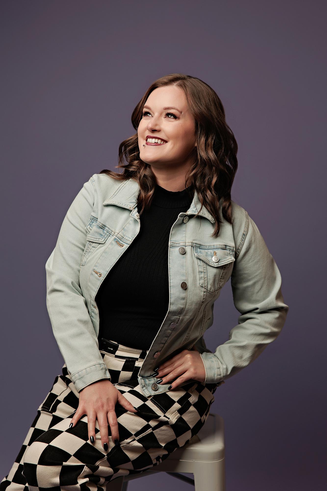 Fun personal branding photography Calgary studio: Joyful young woman in a light denim jacket and checker pants seated on a stool laughing upward, purple background