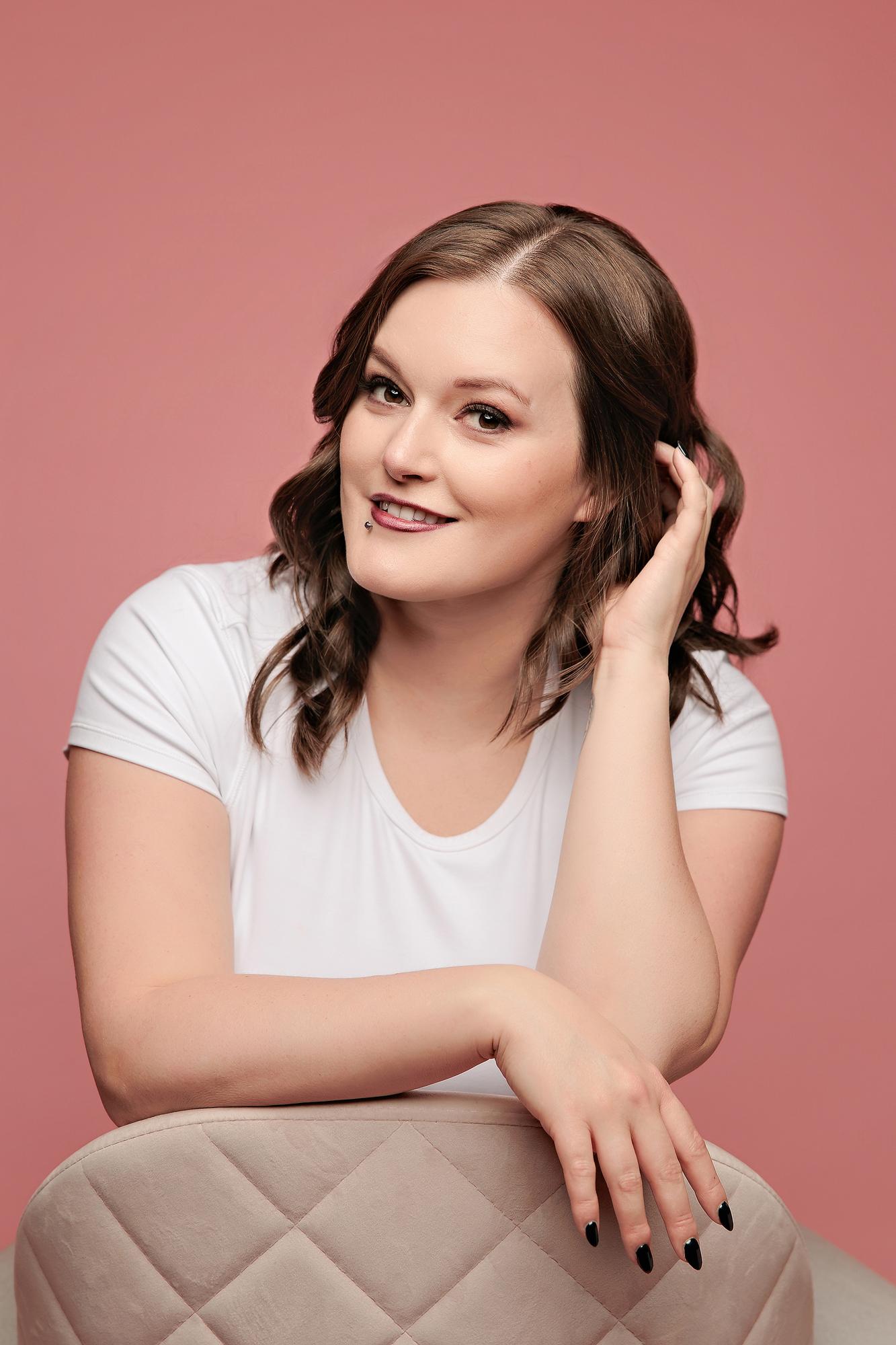 Lifestyle personal branding headshot Calgary: Relaxed young woman in a white t-shirt leaning on a quilted beige chair with hand in hair, warm pink studio background