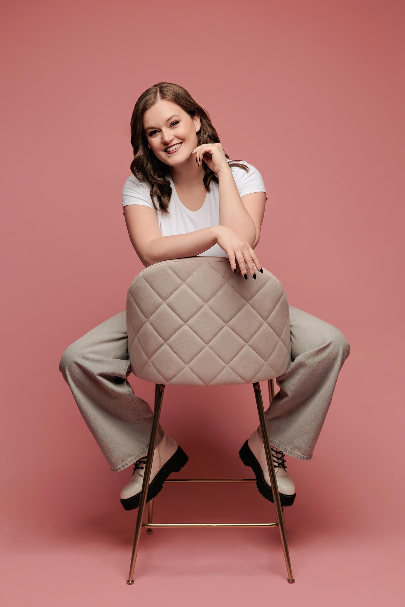 Creative personal branding photography Calgary: Smiling young woman in a white t-shirt and wide-leg grey jeans straddling a quilted chair with chin resting on hands, pink background