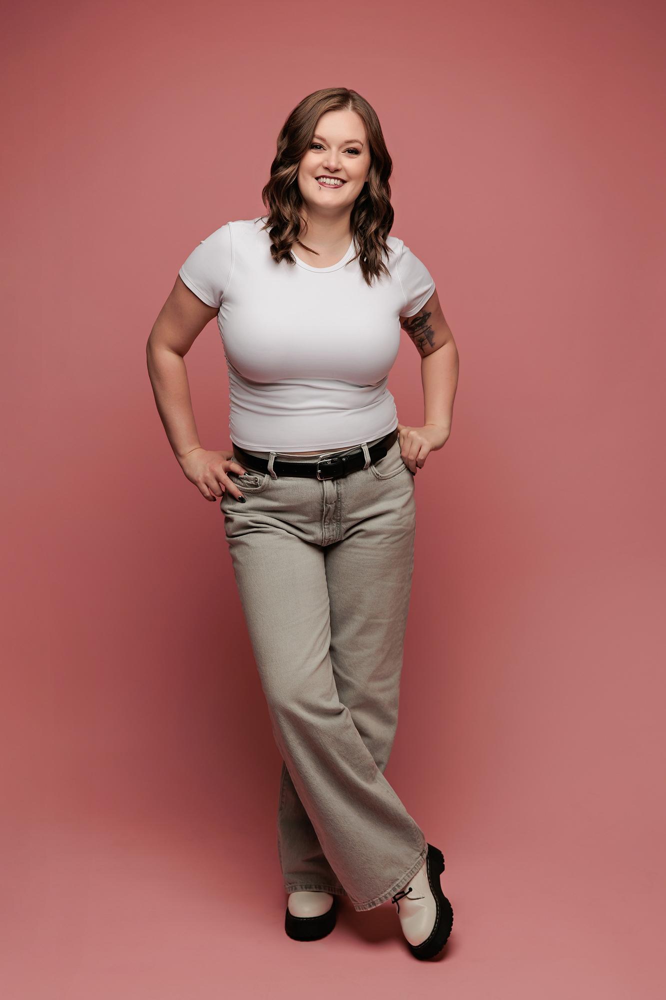 Full-length personal branding portrait Calgary: Confident young woman in a white t-shirt and wide-leg grey jeans standing with hands on hips against a warm pink studio background