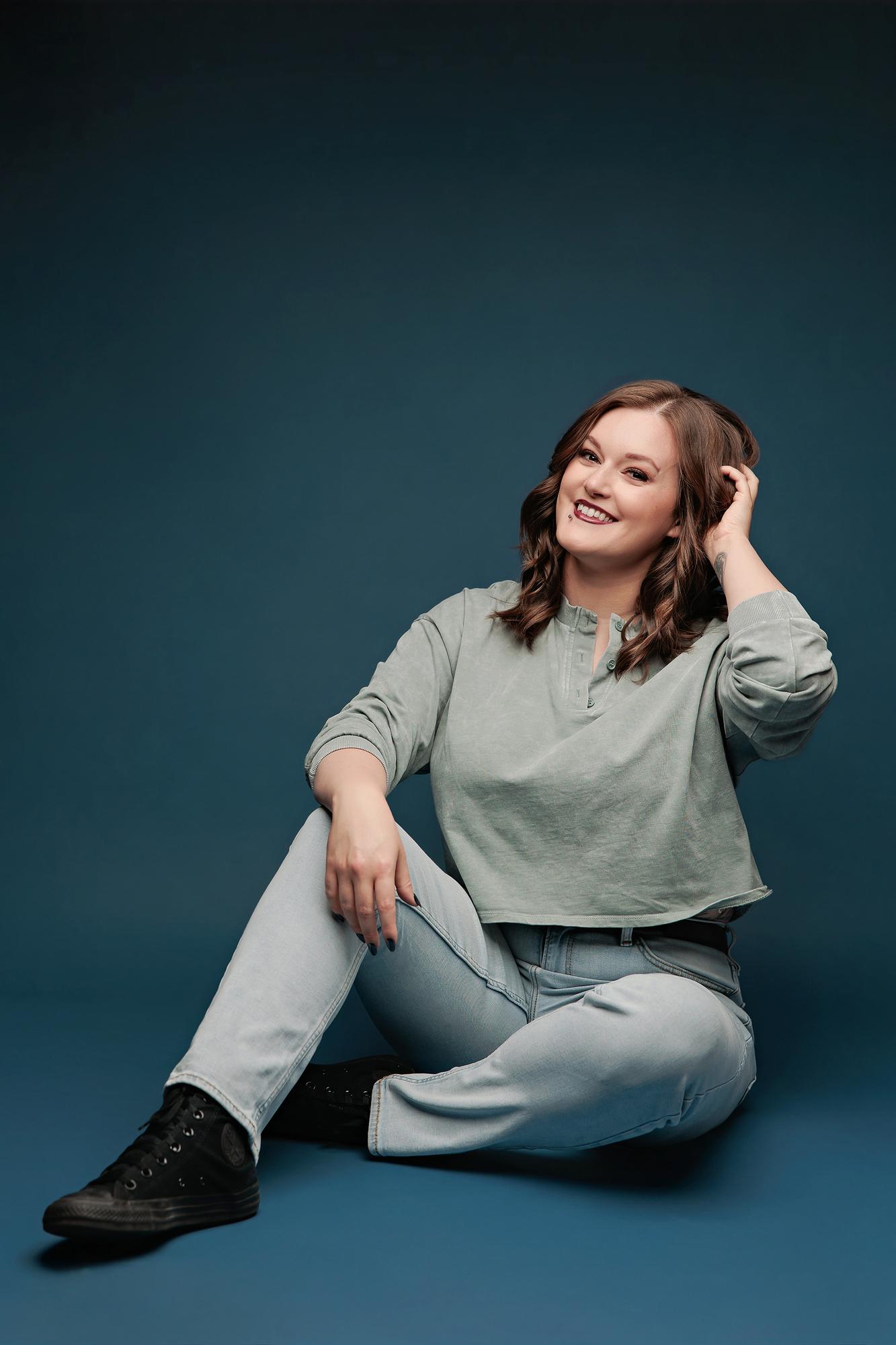 Fun personal branding photography Calgary: Joyful young woman in a sage green top seated on the floor laughing with hand in hair against a teal studio background