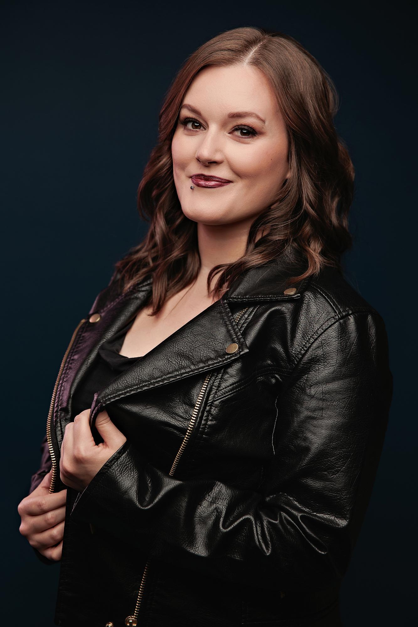 Edgy personal branding headshot Calgary: Young woman in a black leather moto jacket posing confidently in three-quarter profile against a deep teal background
