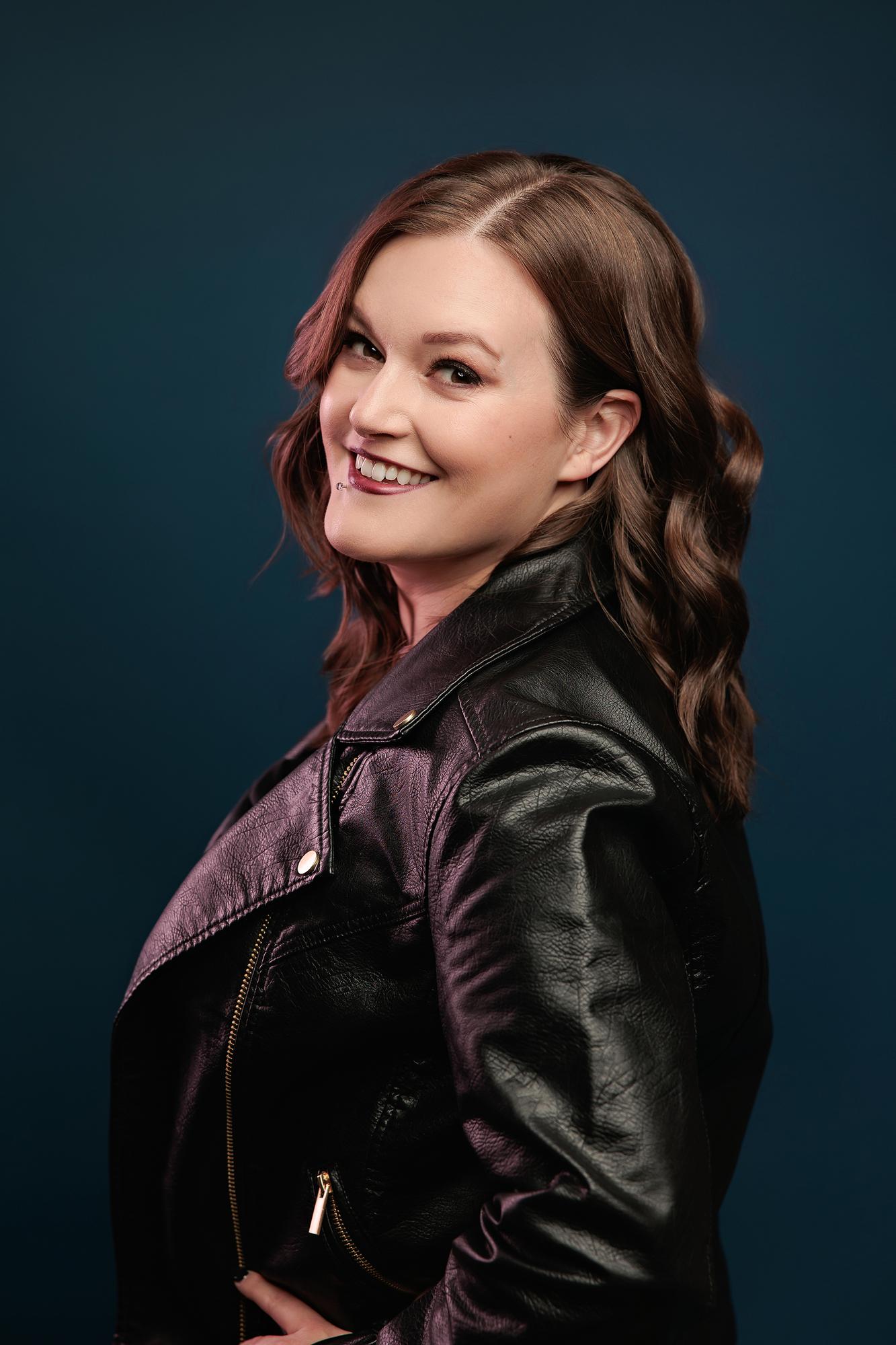 Creative branding portrait Calgary studio: Laughing young woman in a black and purple leather jacket glancing over her shoulder against a deep teal background