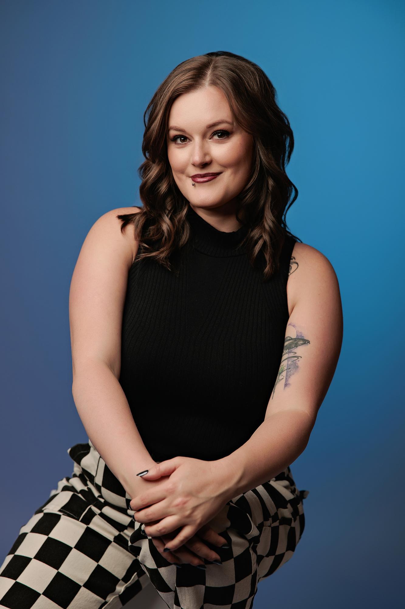 Modern personal branding photography Calgary: Young woman in a black sleeveless ribbed top and bold black and white checker pants seated with hands clasped, bright blue background