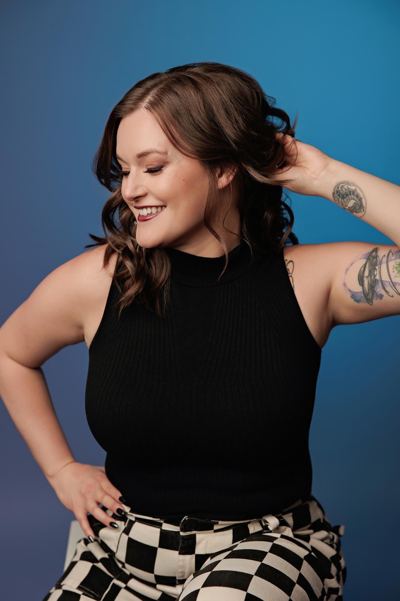 Lifestyle personal branding Calgary studio: Young woman in a black top and checker pants smiling downward with hand in hair against a bright blue backdrop