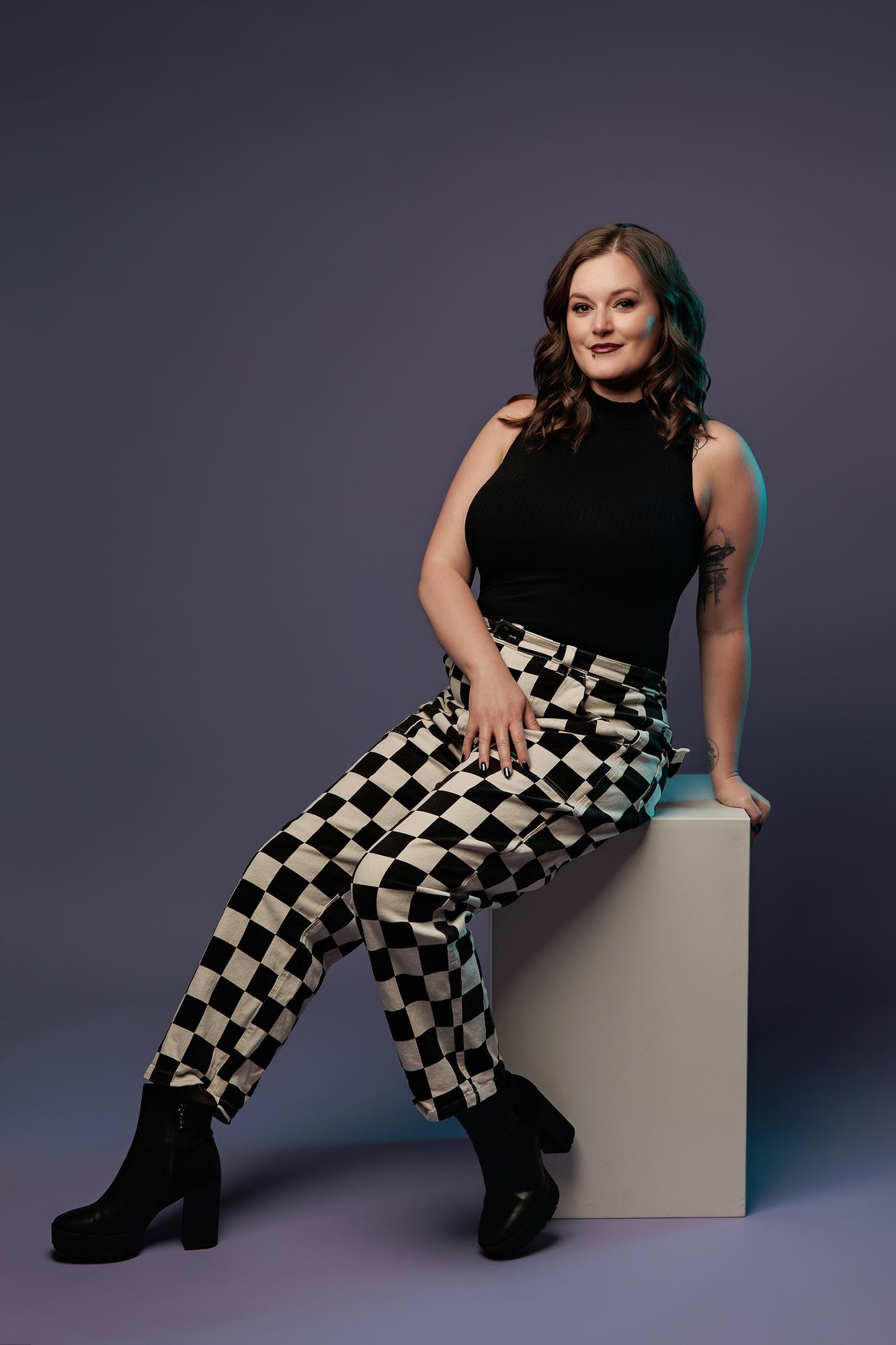 Editorial personal branding photography Calgary: Young woman in a black sleeveless top and checker pants leaning on a white cube with a teal light accent, purple background