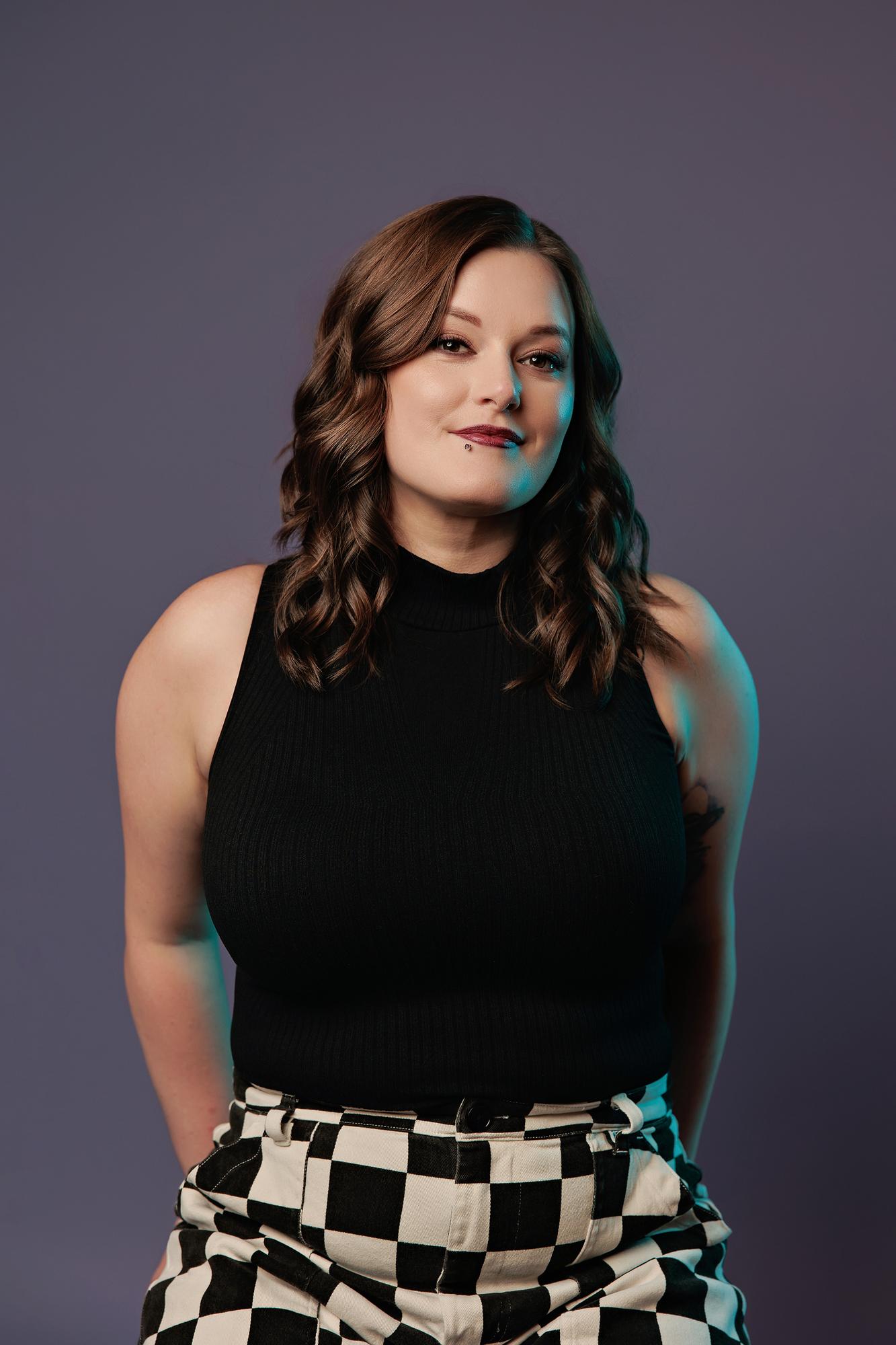 Bold personal branding photography Calgary: Young woman in a black sleeveless top and checker pants standing confidently with a teal light accent against a purple background