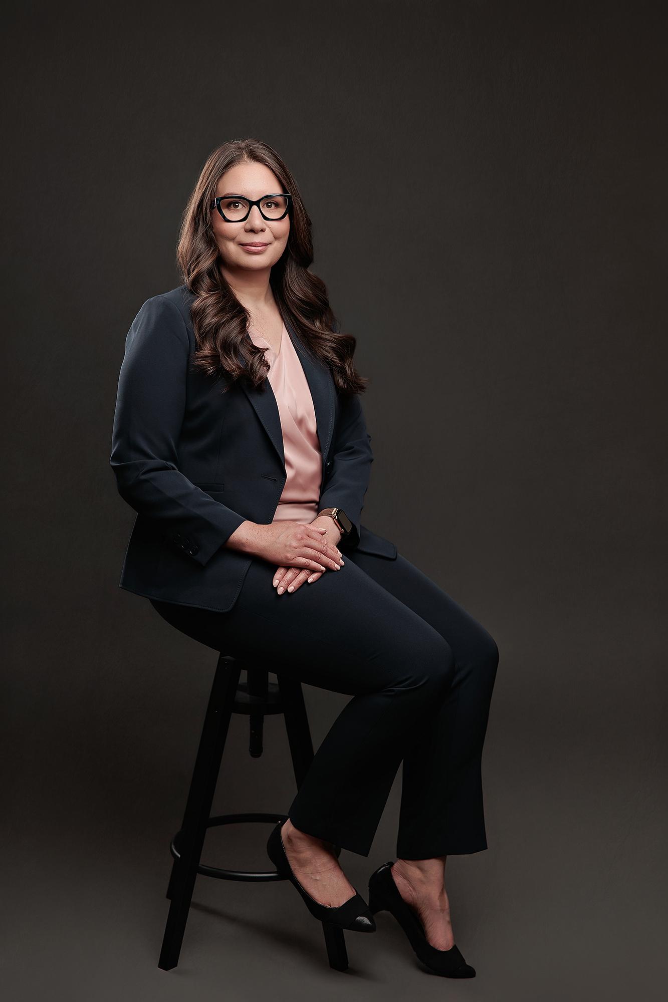 A professional woman in a navy suit sits on a stool against a dark backdrop, showcasing the polished style of our Calgary headshot photography studio.