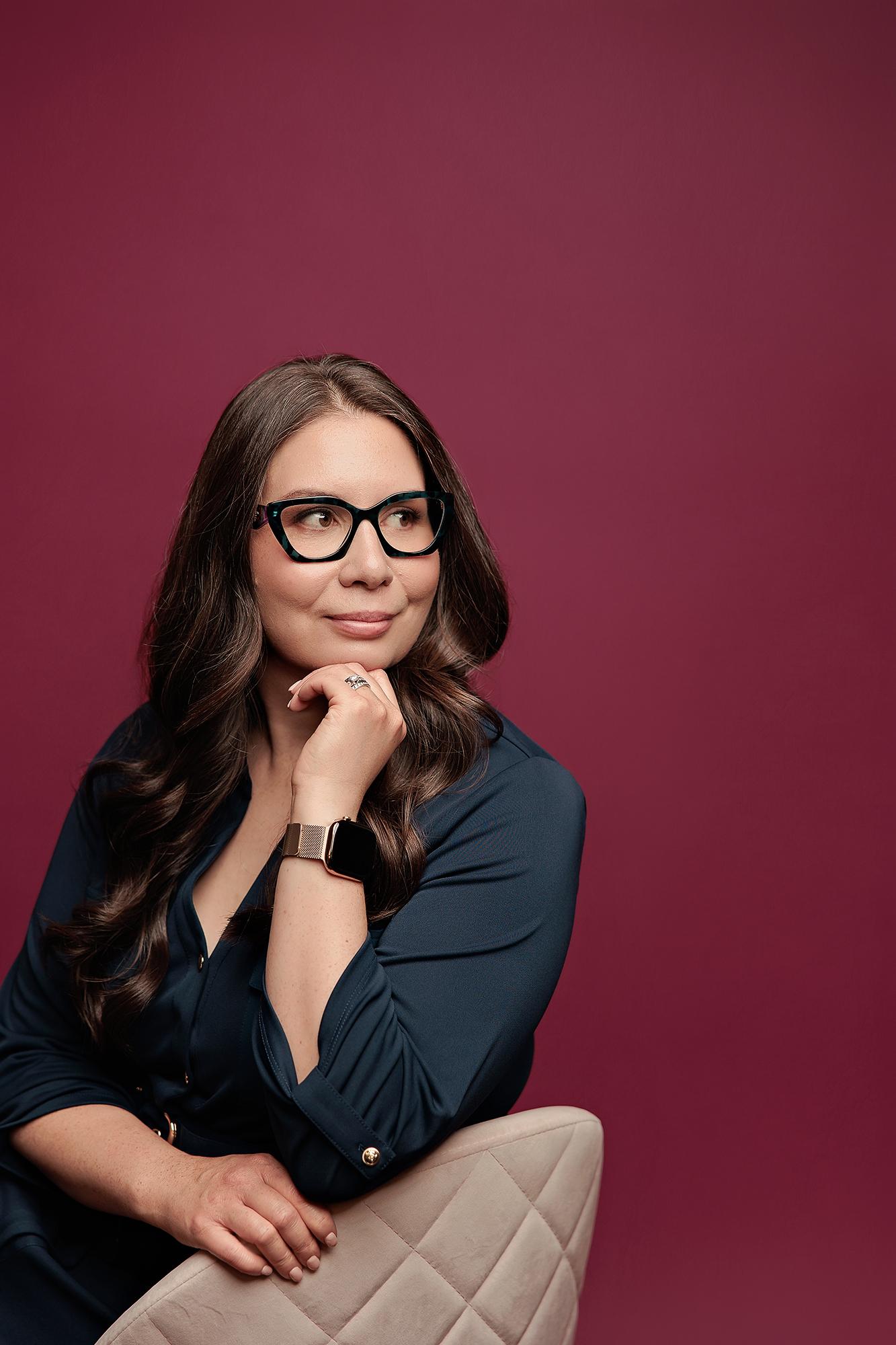 A professional woman in a dark blouse poses thoughtfully for this Calgary headshot, featuring soft studio lighting against a bold burgundy backdrop.