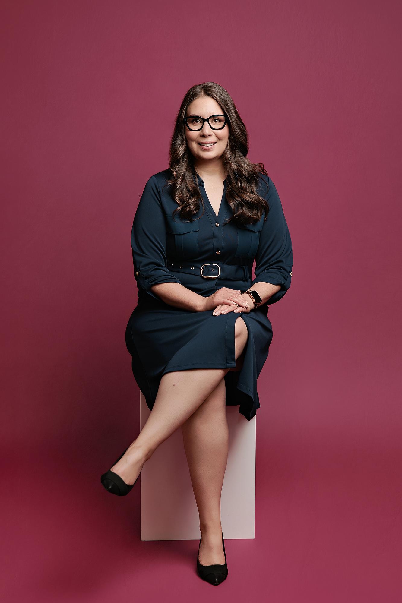 A professional woman in a dark teal dress sits on a stool against a deep red backdrop, showcasing the polished style of a Calgary headshot photographer.