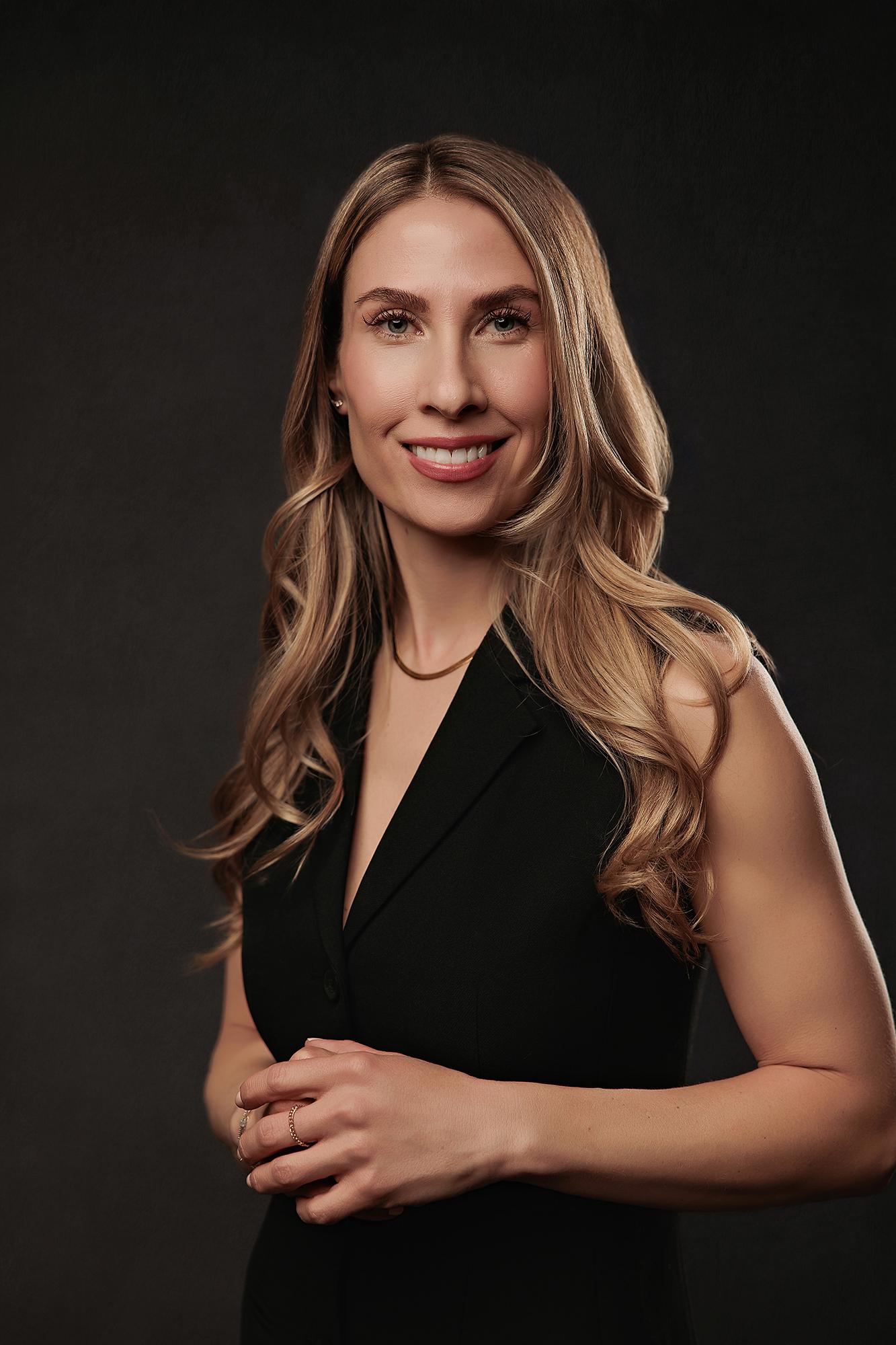 Professional headshot Calgary studio: Close-up portrait of a woman with long blonde-highlighted wavy hair in a black sleeveless vest dress with hands clasped and a warm smile, gold collar necklace visible, dark charcoal background