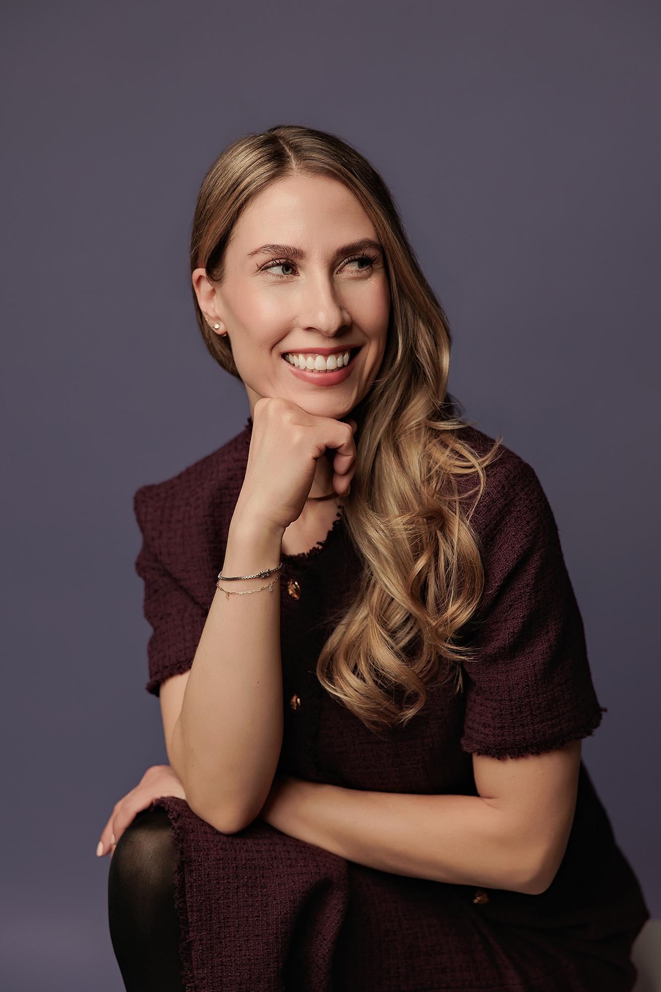 Professional headshot Calgary studio: Joyful woman with long blonde-highlighted wavy hair in a burgundy tweed dress, close-up with chin resting on fist and a radiant upward smile, delicate layered bracelets visible, soft purple background