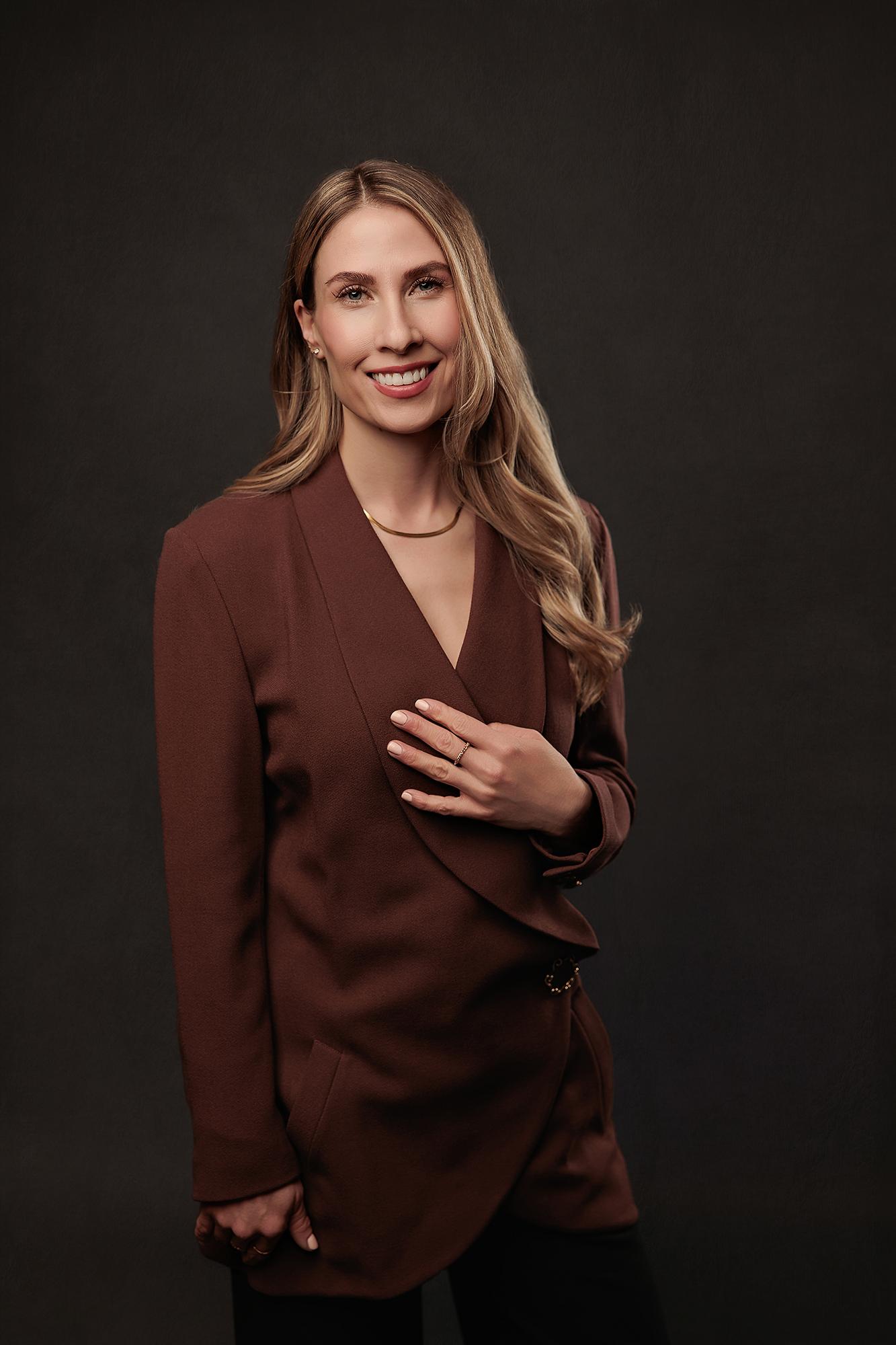Professional headshot Calgary studio: Confident woman with long blonde-highlighted wavy hair in a chocolate brown wrap blazer with a gold collar necklace, hand resting on her chest and a warm smile against a dark charcoal background