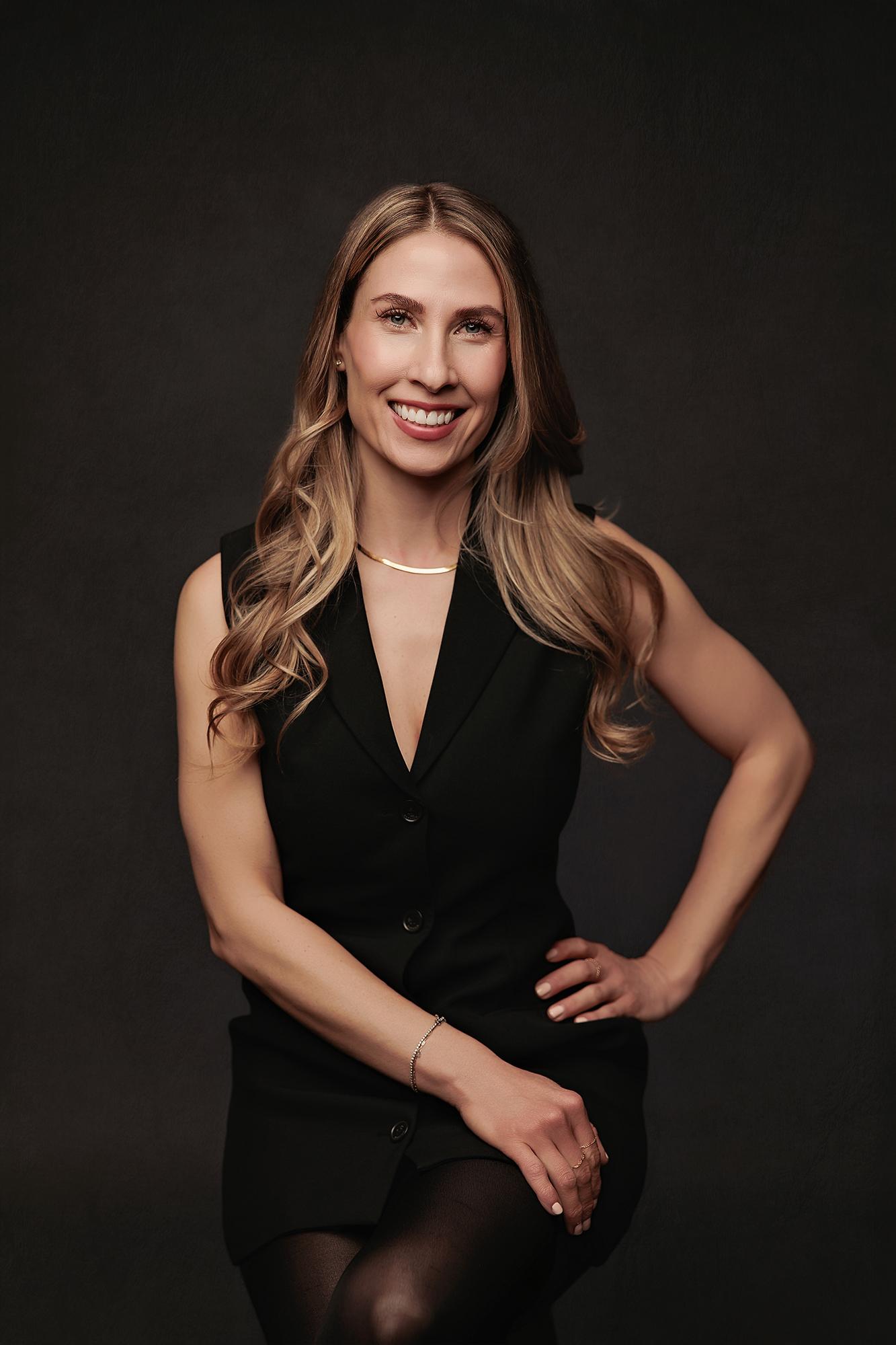 Professional headshot Calgary studio: Powerful woman with long blonde-highlighted wavy hair in a black sleeveless button-front vest dress with hand on hip, gold collar necklace and delicate bracelet, smiling brightly against a dark charcoal background