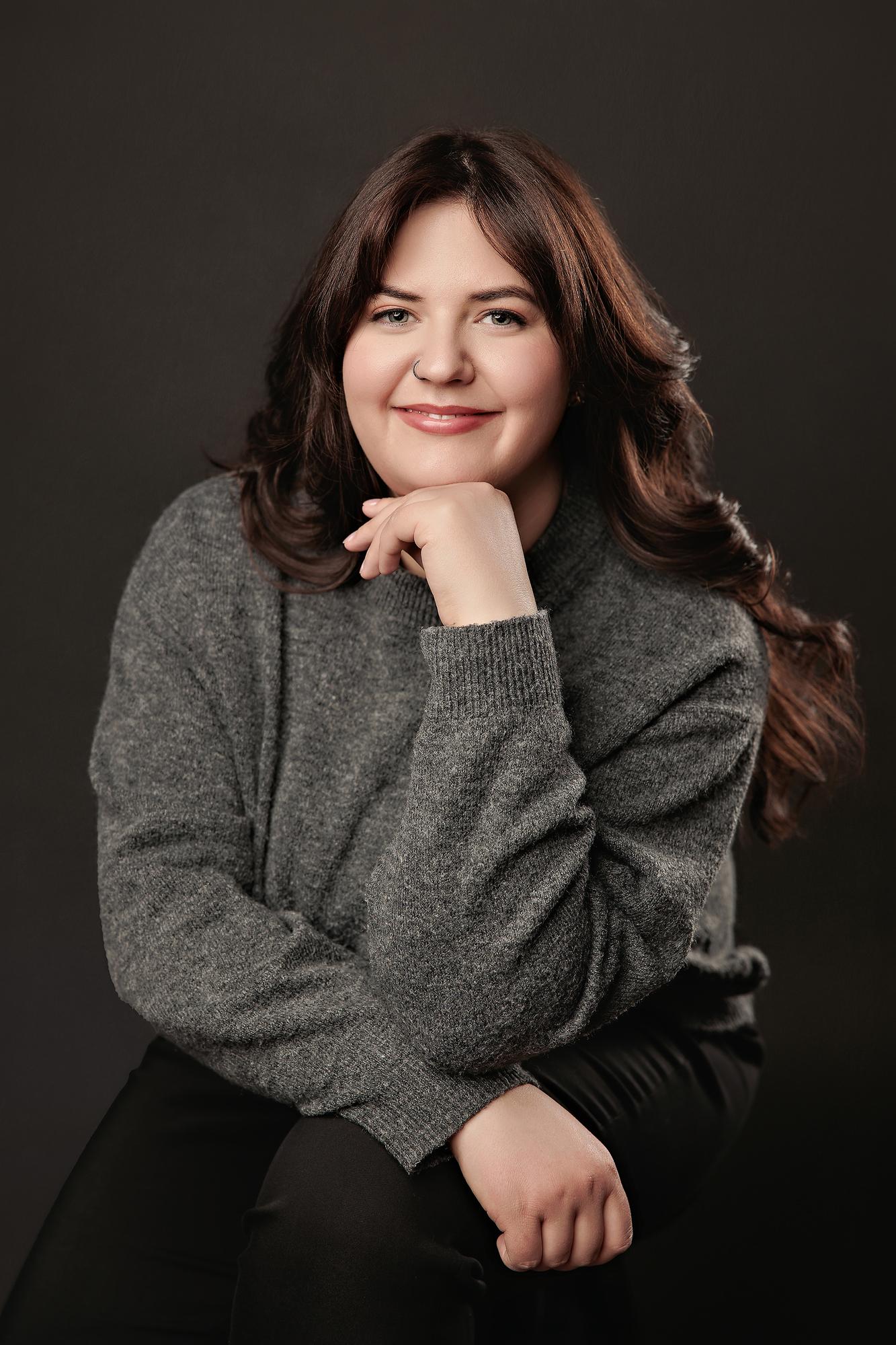 Professional headshot Calgary studio: Warm and approachable young woman in a charcoal grey oversized sweater resting her chin on her hand against a dark background