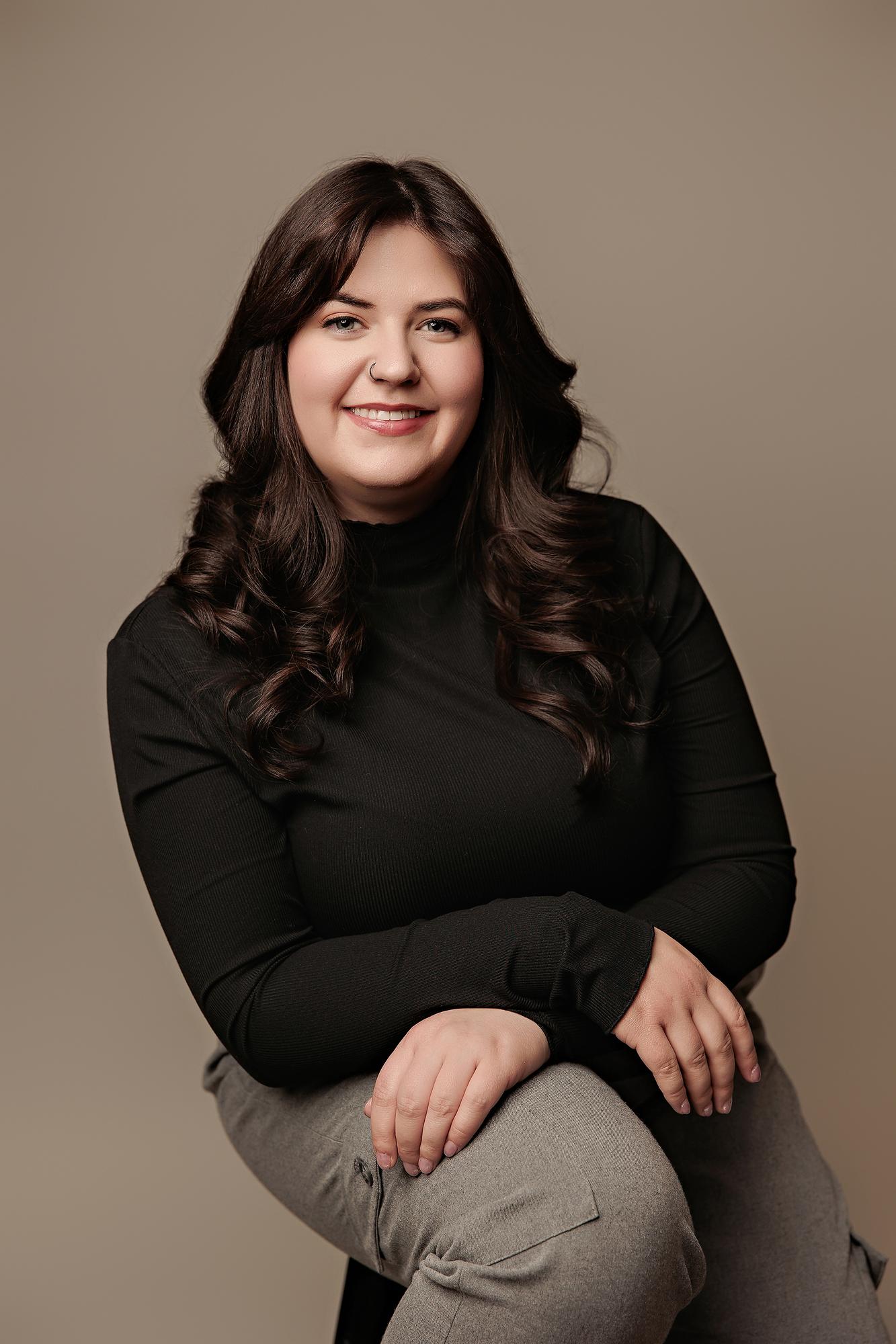 Corporate headshot photography Calgary: Smiling young woman in a black ribbed turtleneck and grey trousers seated in a relaxed pose against a warm taupe studio background
