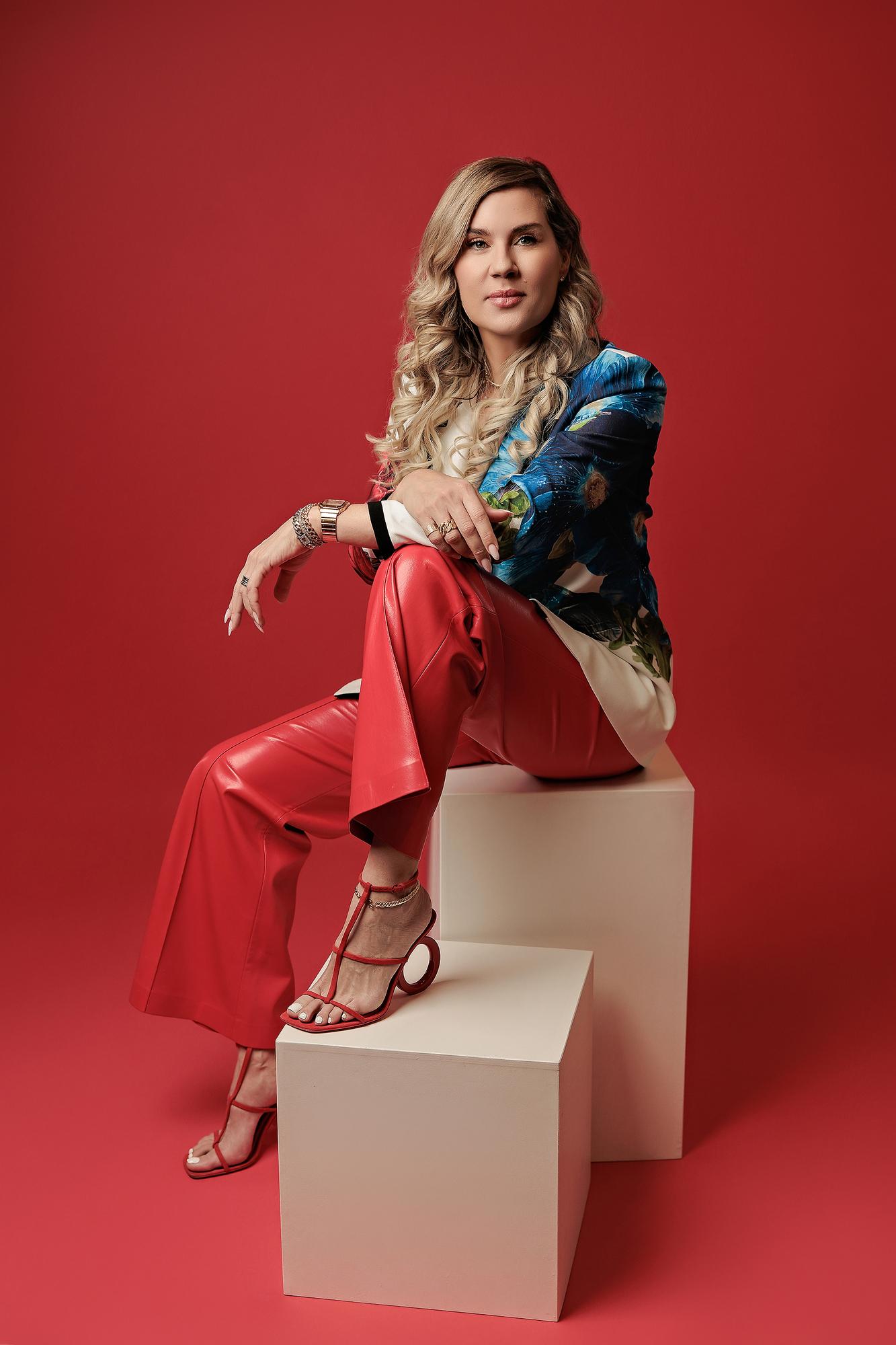 Fashion-forward professional headshot Calgary: Woman in a bold floral blazer and wide-leg red leather pants seated on white cubes with red heeled sandals against a matching red background