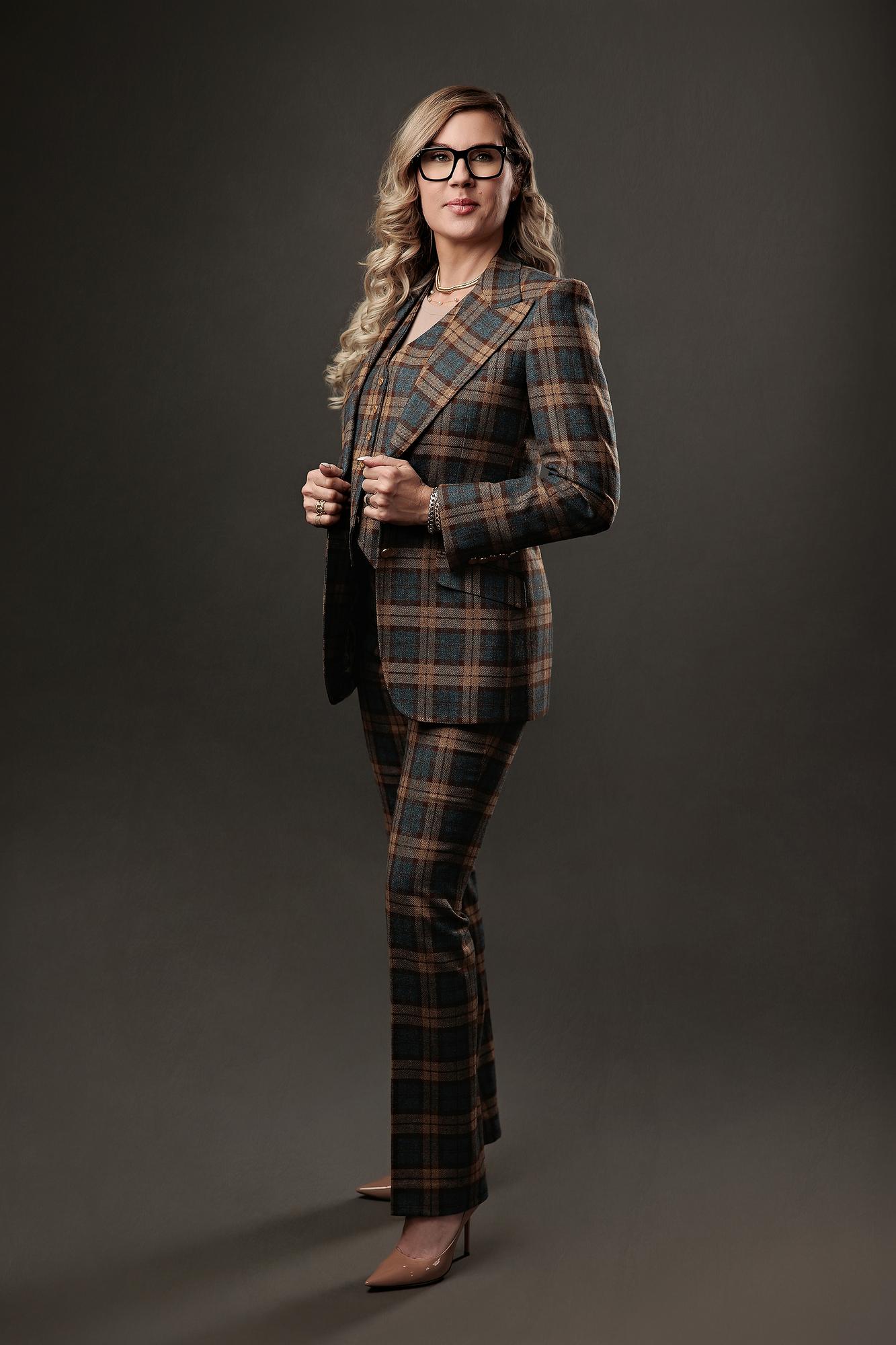 Full-length professional headshot Calgary: Striking woman in a brown and teal plaid three-piece suit and black-frame glasses standing confidently against a dark studio background