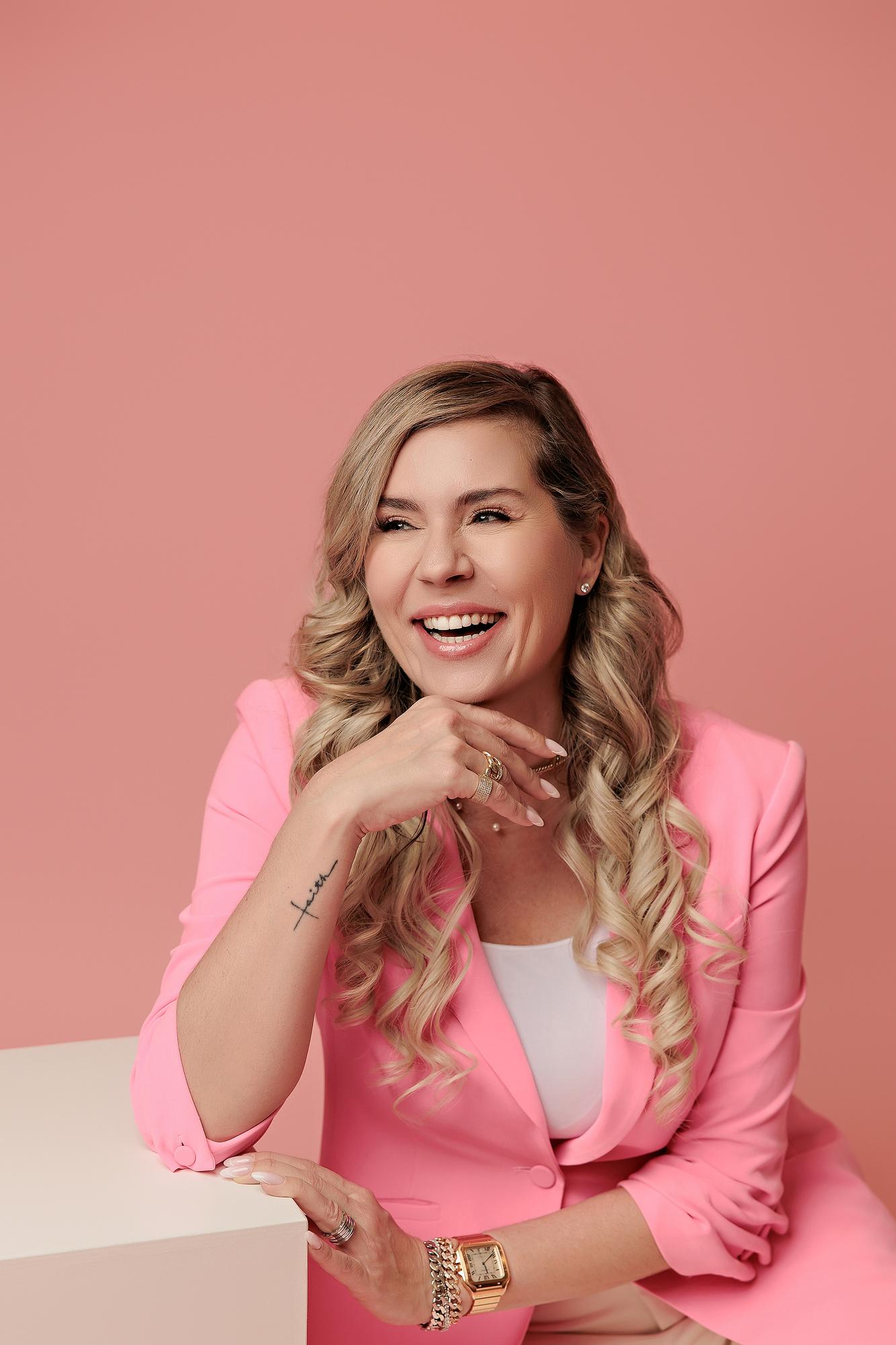Vibrant personal branding headshot Calgary: Joyful woman in a hot pink blazer and white top laughing while leaning on a white cube against a warm pink studio background
